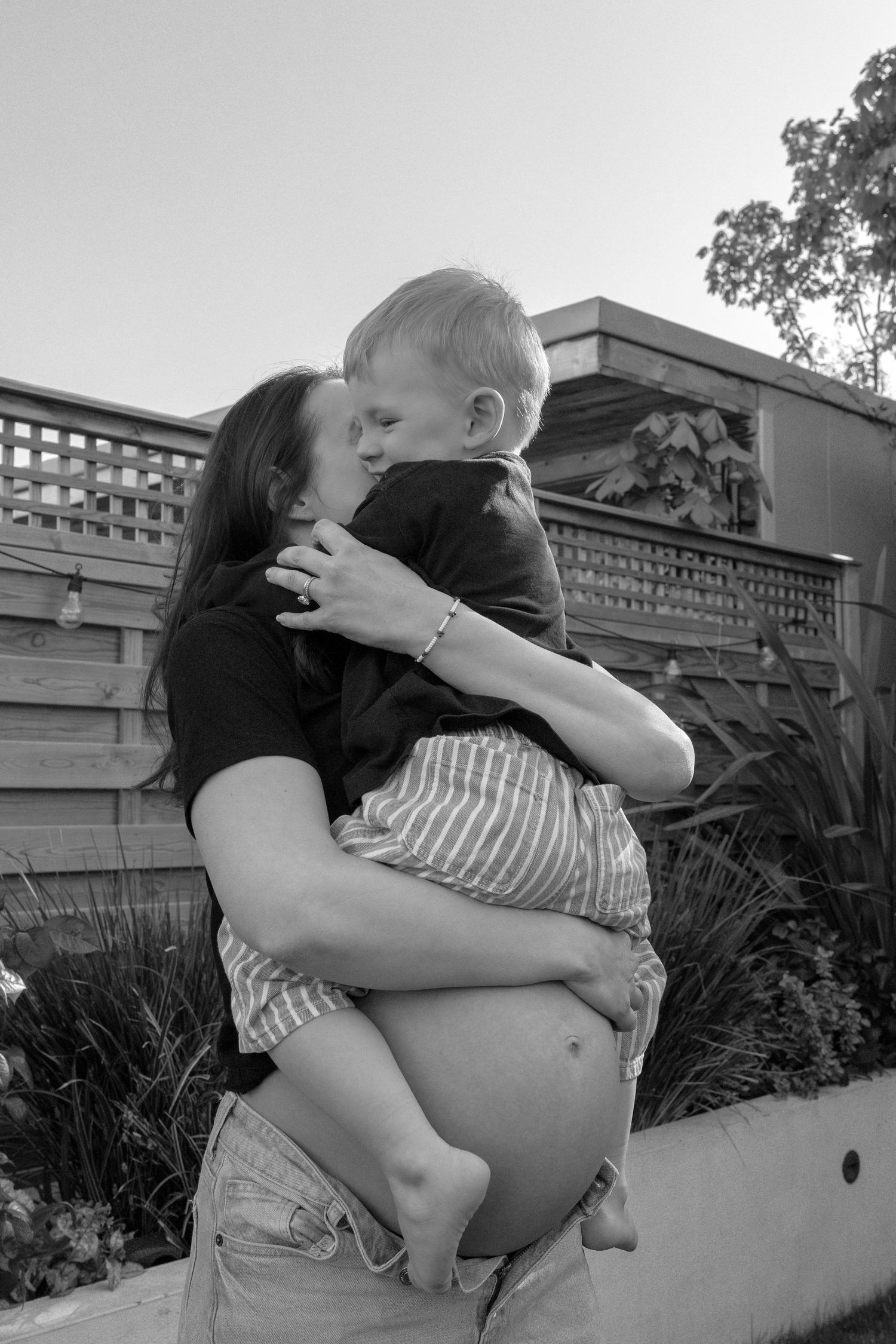 Pregnant woman in black top holds young blonde boy who kisses her cheek in a black and white garden photo.