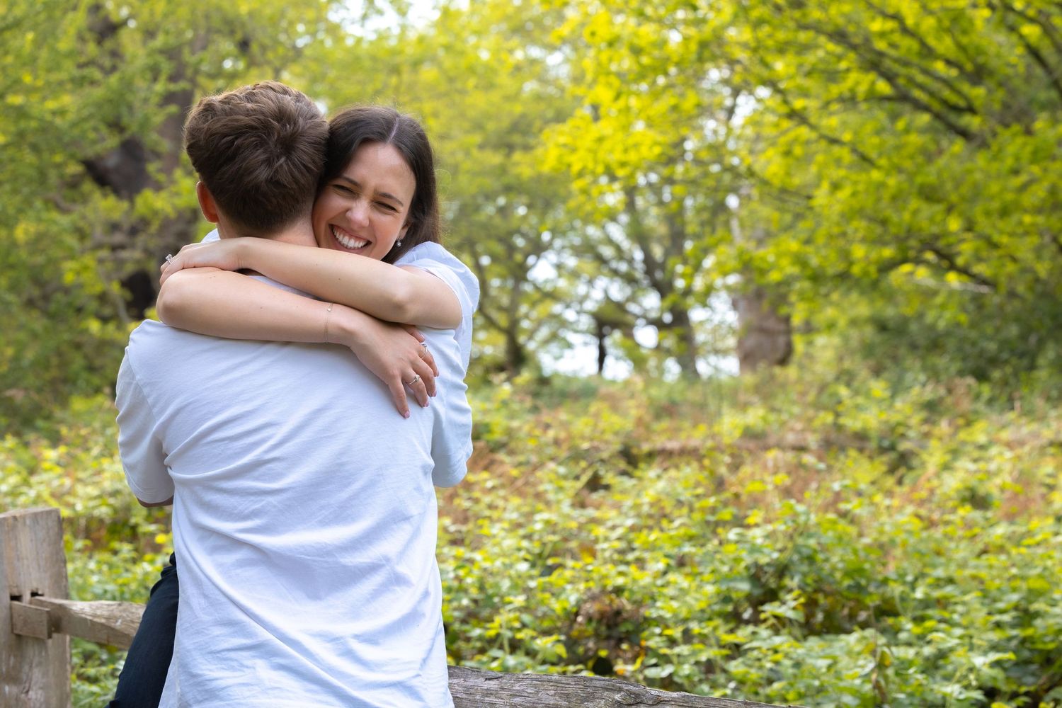 A smiling woman hugs her partner outdoors surrounded by autumn green trees.
