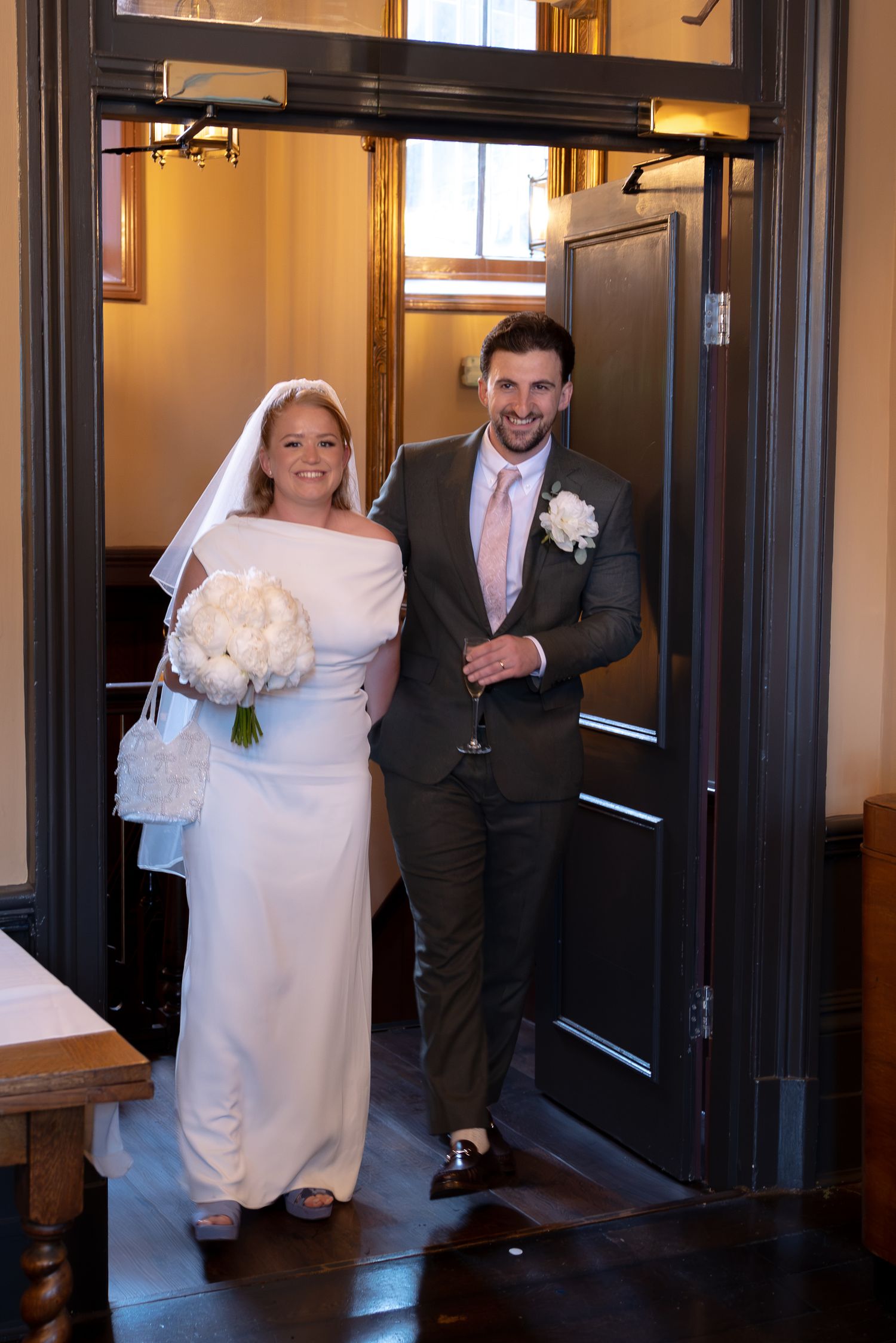 Smiling bride in white gown and groom in grey suit entering a venue doorway together.