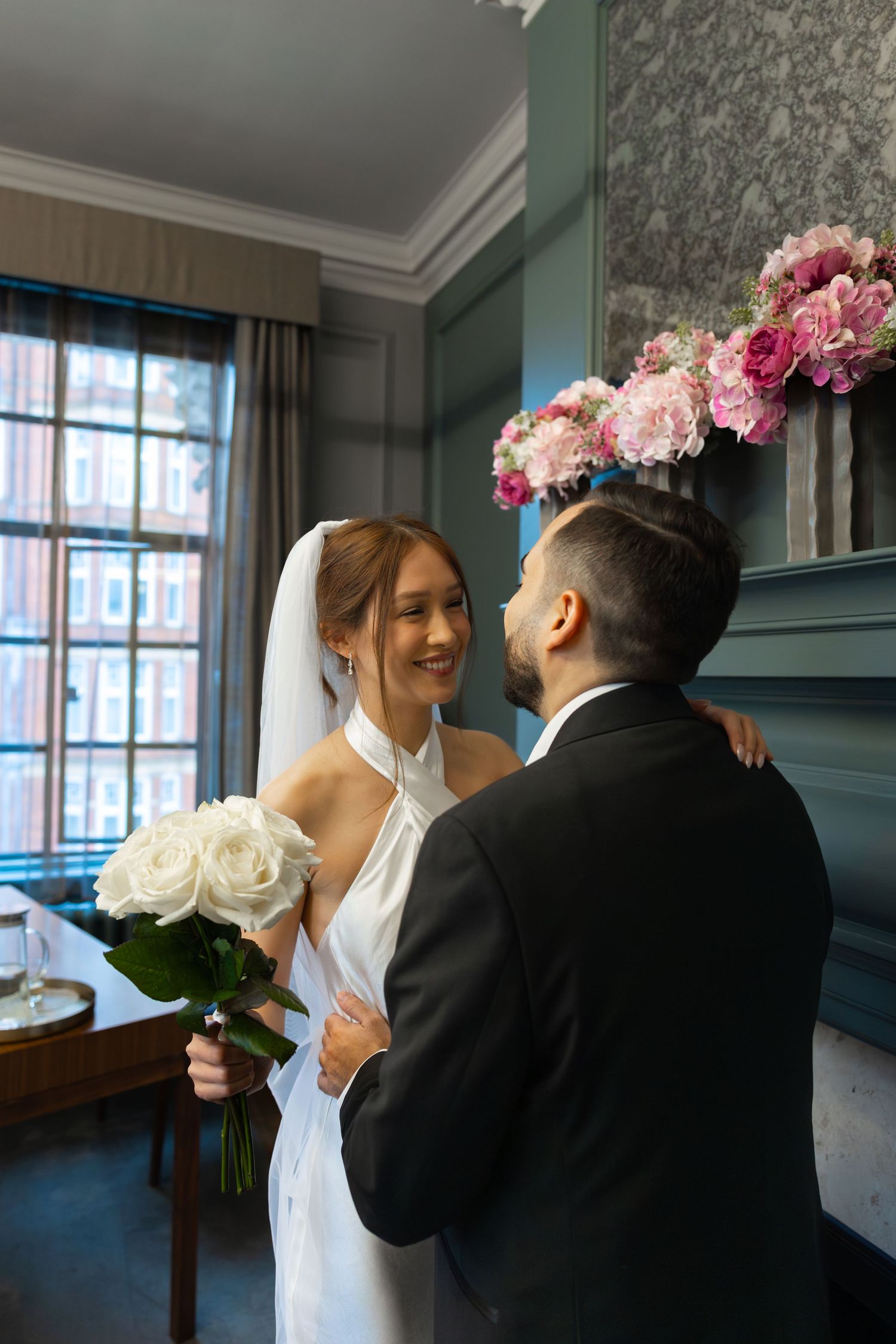 Smiling bride in white dress and veil holding roses, embracing groom in black suit in elegant room.