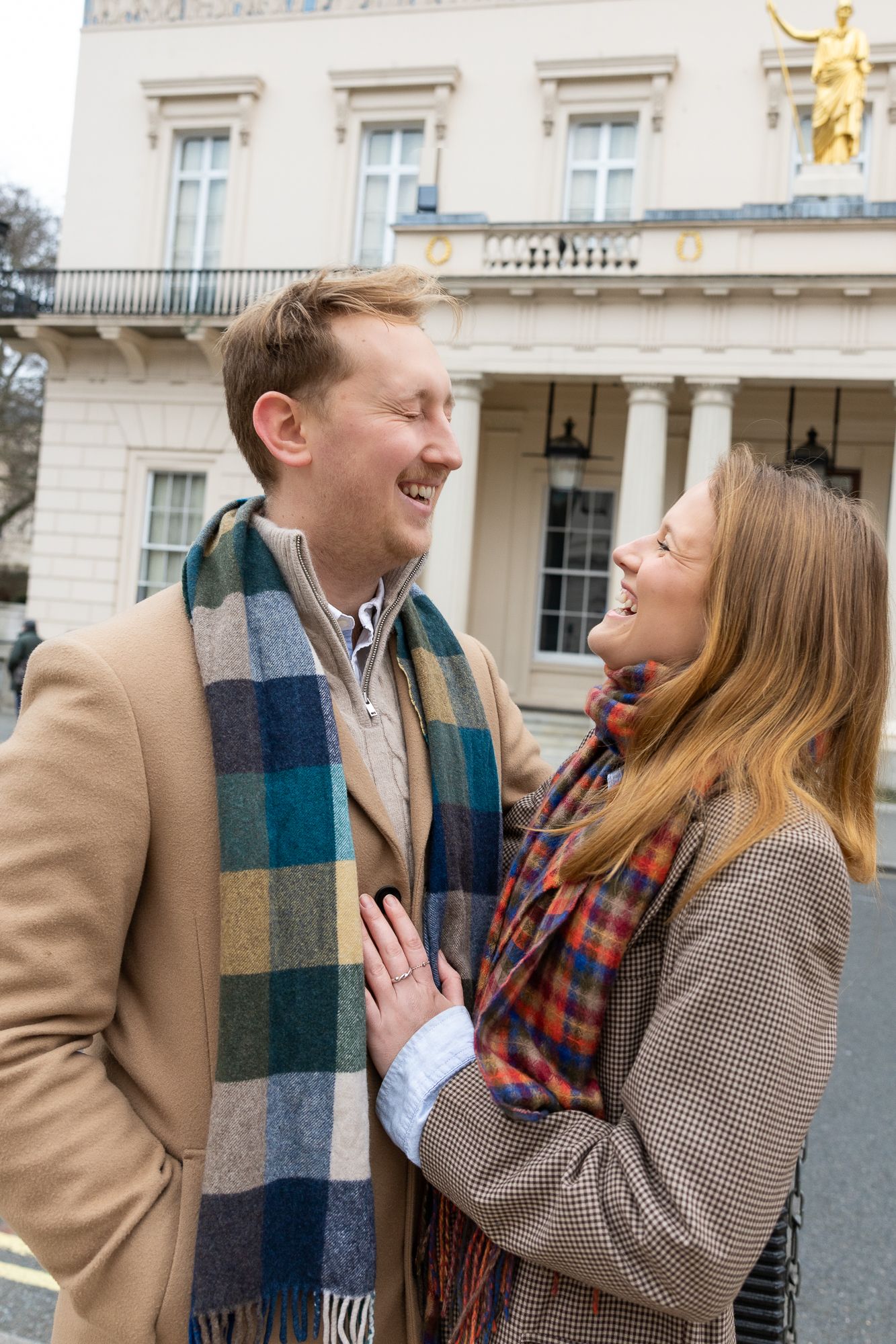 Two people share an intimate moment of laughter outside an elegant building with golden statues.