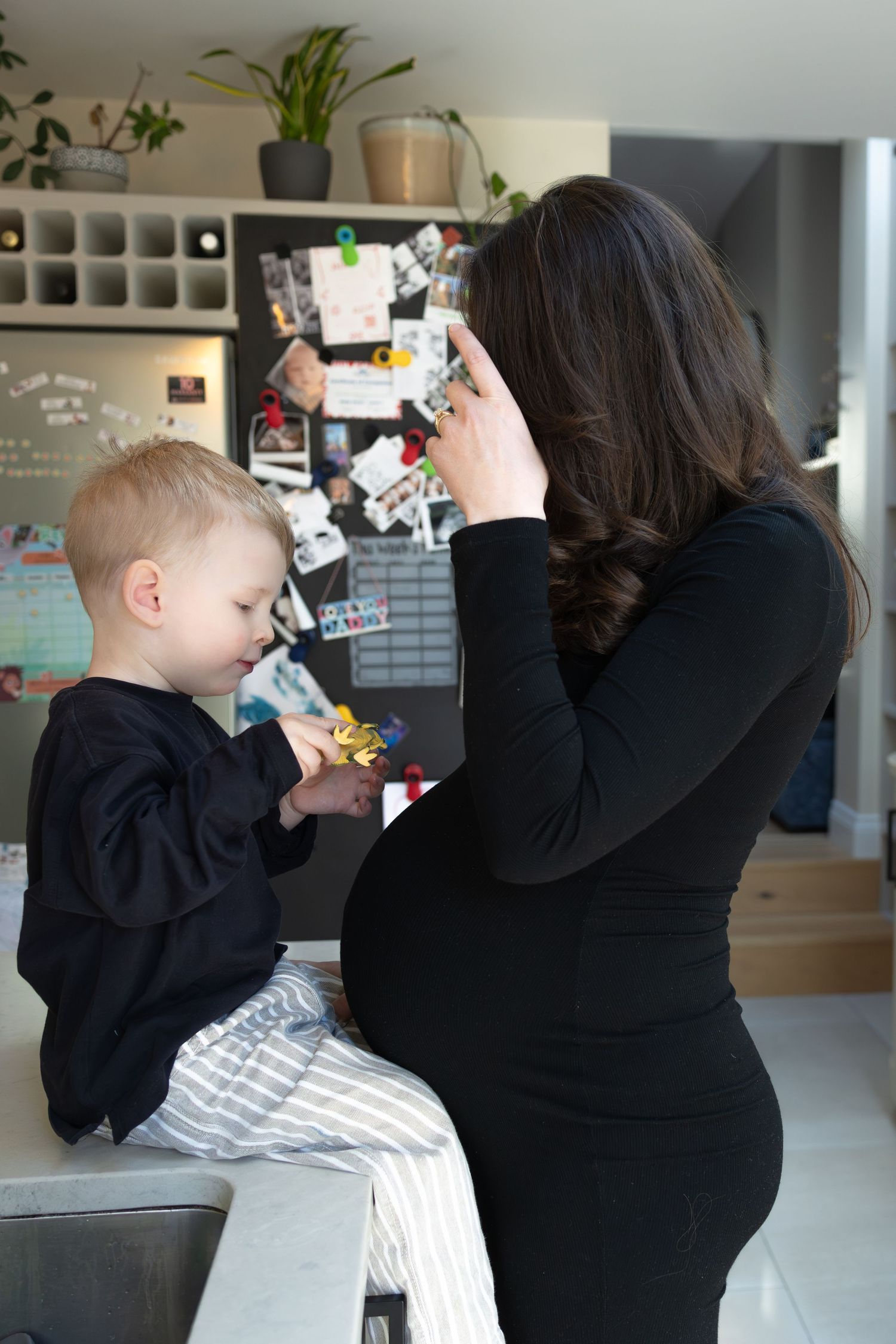 Pregnant woman in black dress interacting with toddler son sitting on kitchen counter.