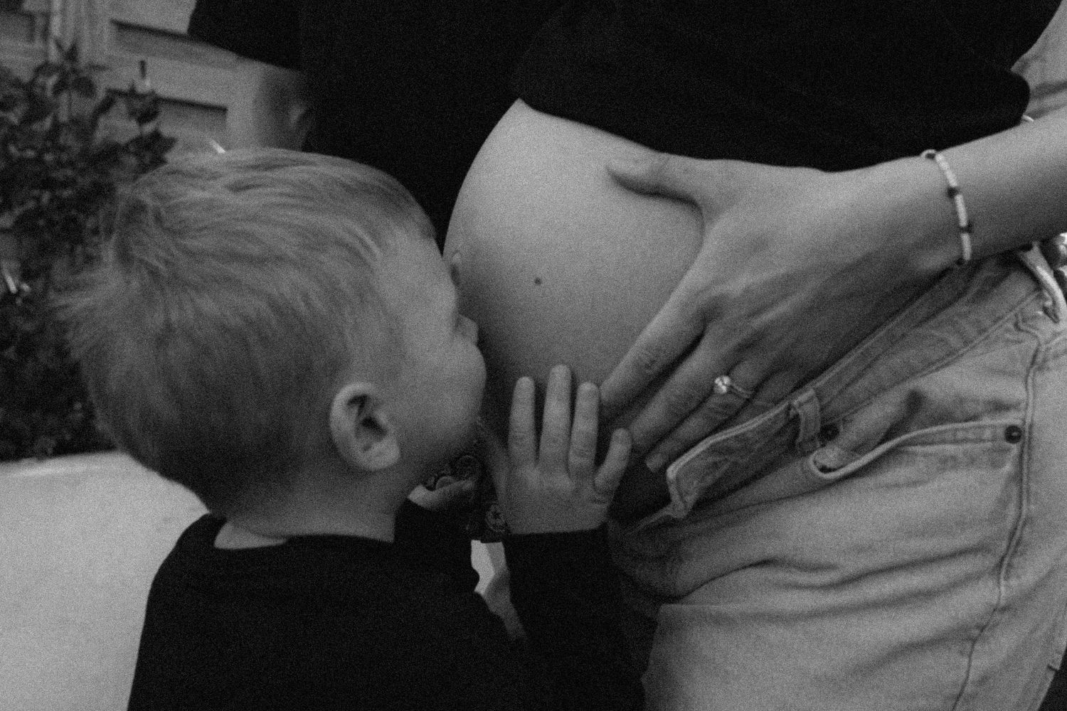 Black and white photo of a young boy kissing his pregnant mothers bare baby bump outdoors.
