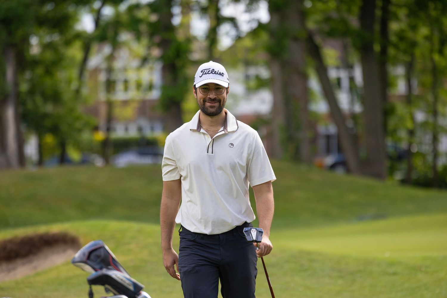 A smiling male golfer in a white polo and dark pants holds a golf club on a lush green course with trees behind.