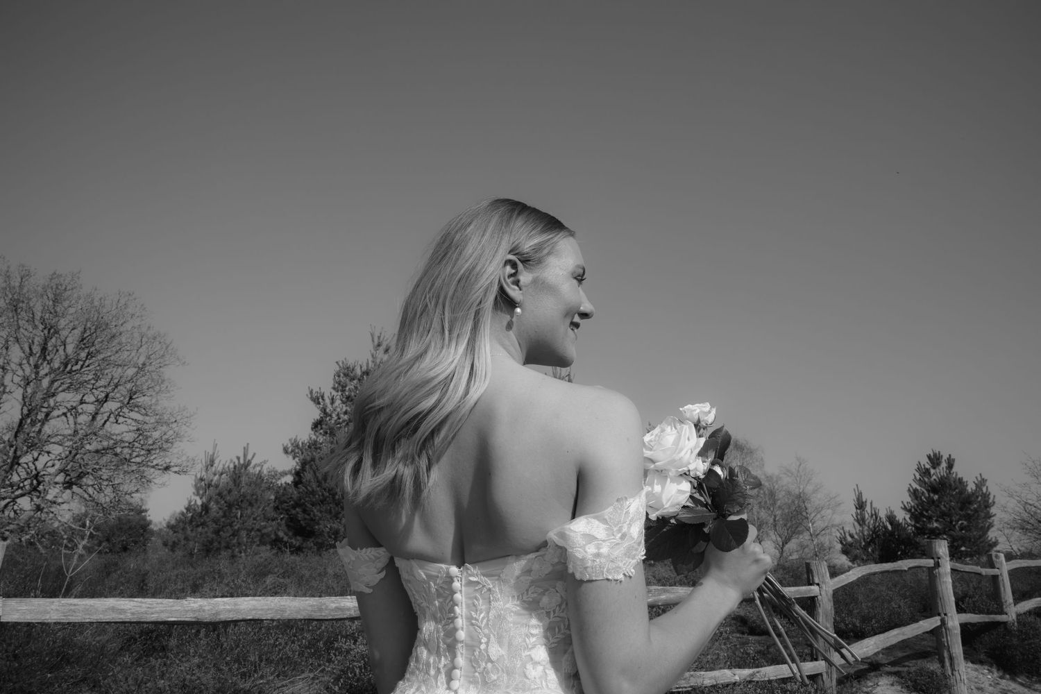 Bride in lace wedding dress holding bouquet, smiling outdoors near wooden fence in black and white photo.