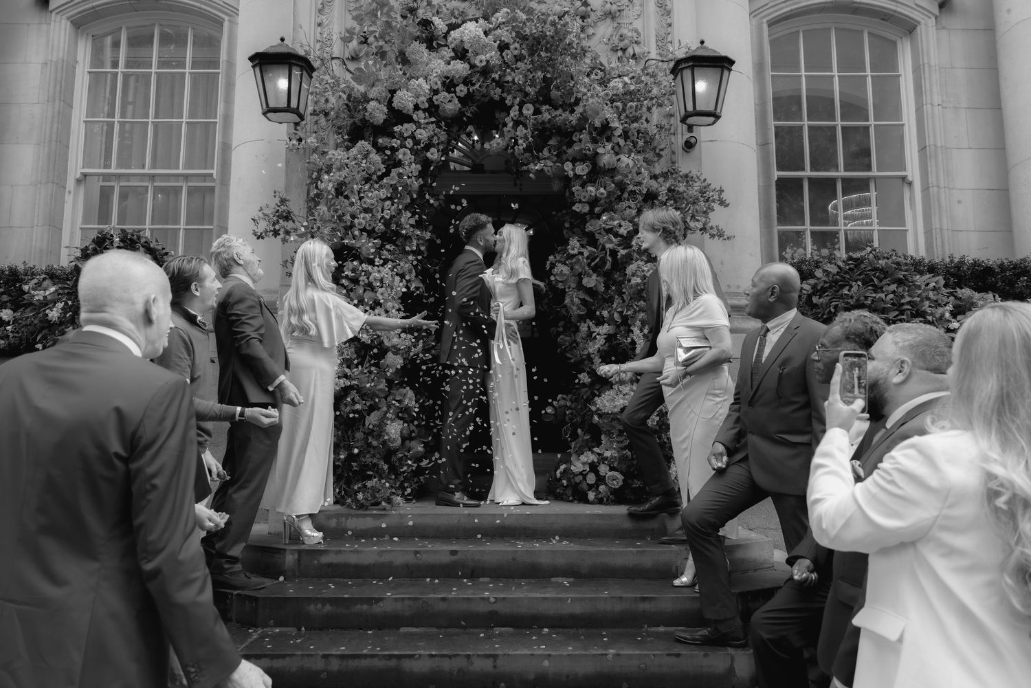Black and white photo of bride and groom on steps surrounded by cheering wedding guests at elegant venue.
