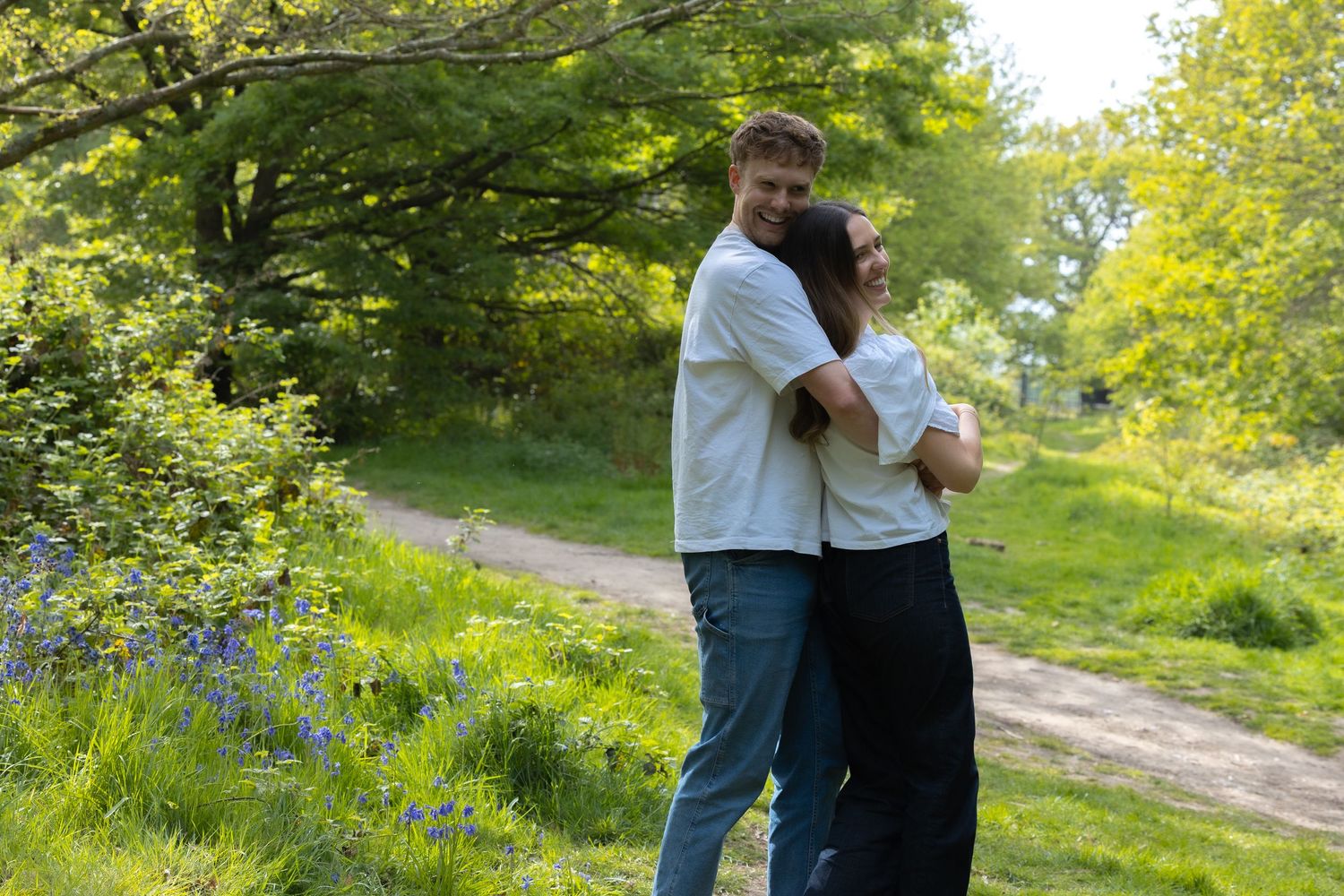 Couple embracing and smiling on a sunny woodland path surrounded by lush green trees and bluebells.