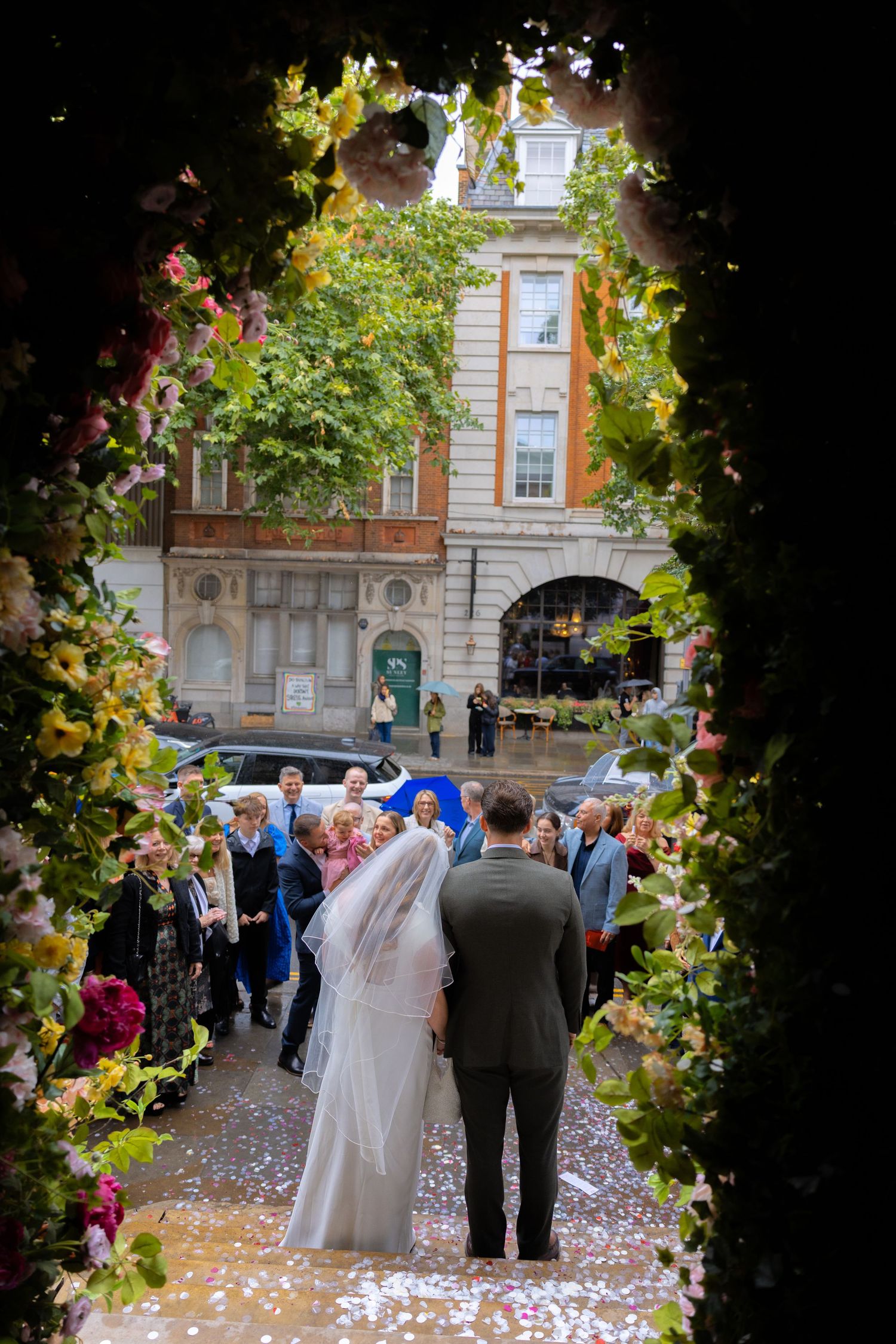 Bride and groom stand at church entrance framed by floral arch with wedding guests and confetti on a London street.