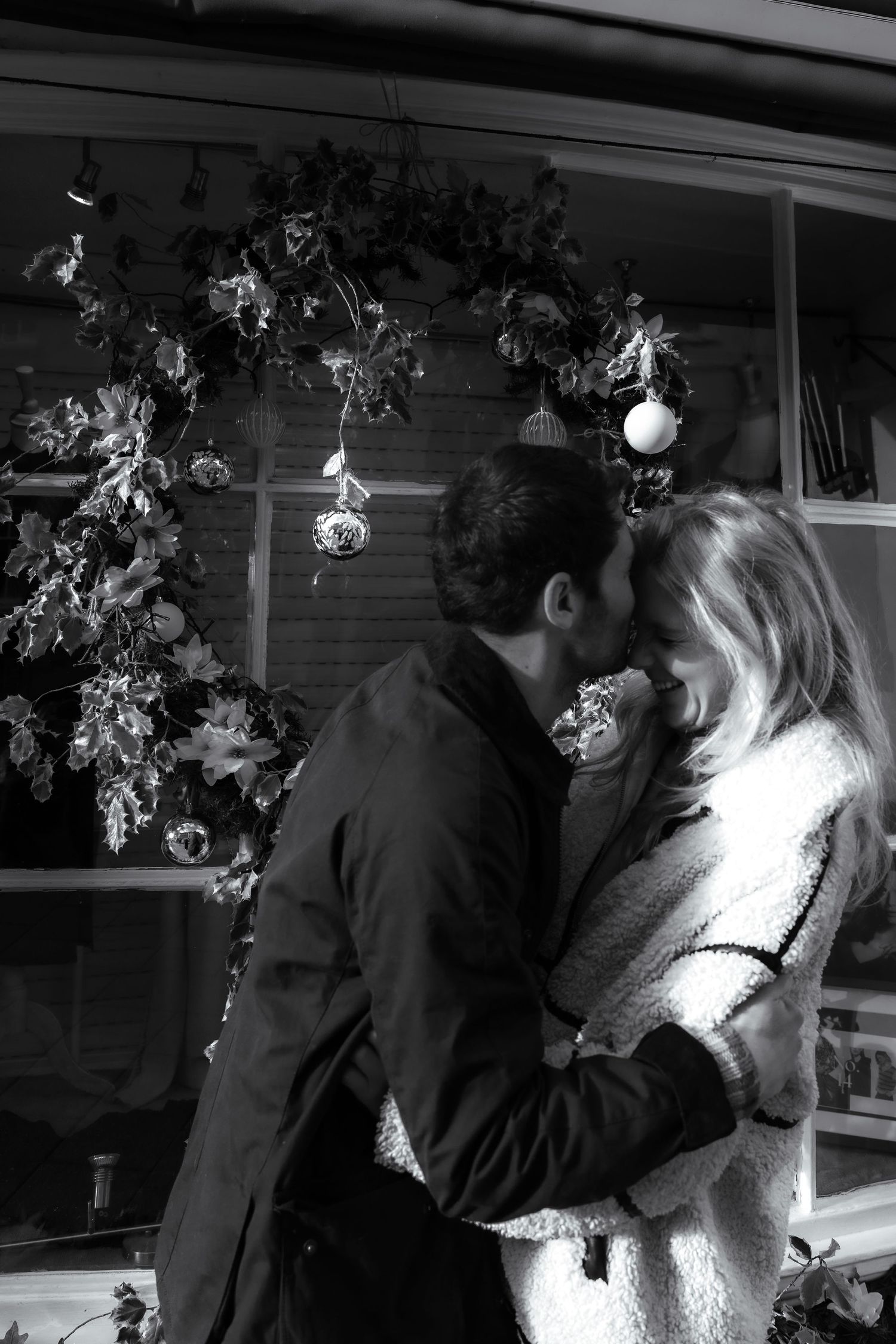 A couple embraces near a window decorated with holiday lights and floral arrangements.