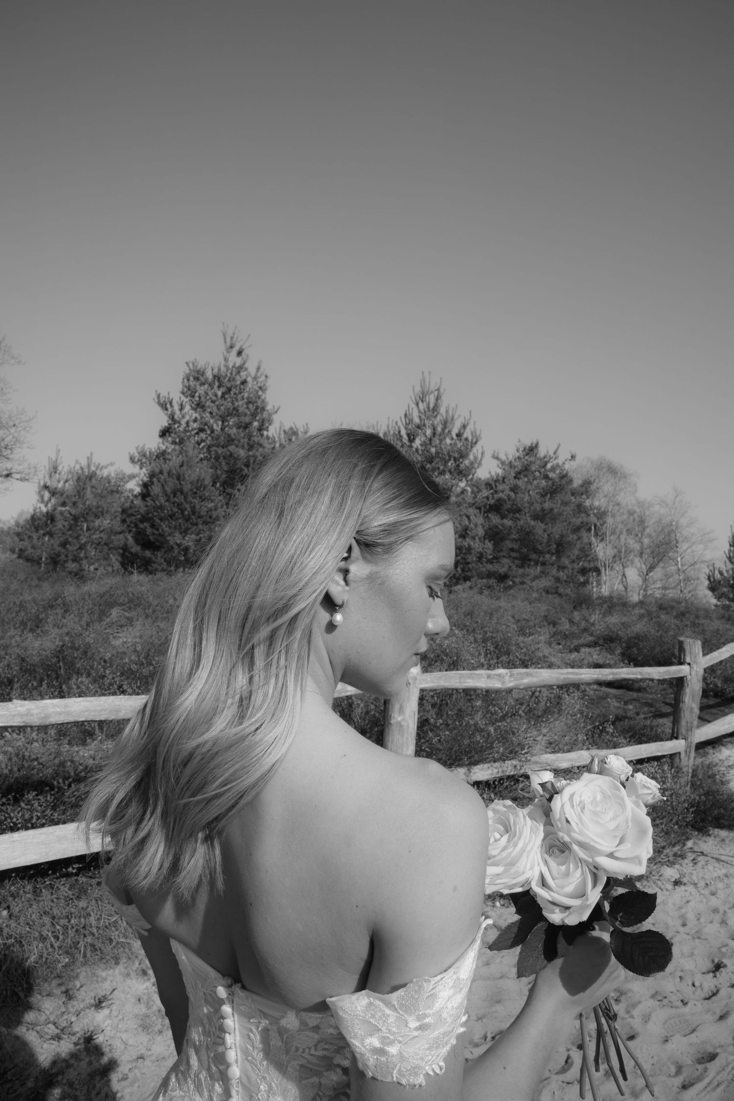 Bride holding white roses bouquet in off-shoulder lace gown, long blonde hair, standing by wooden fence outdoors.
