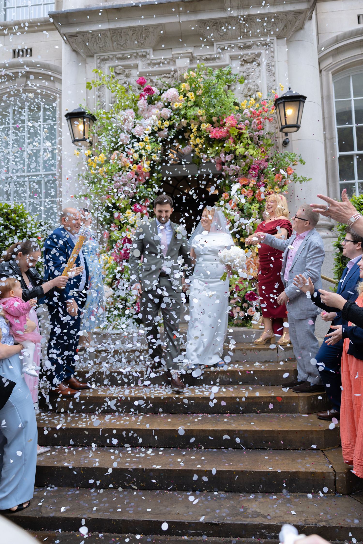Newlyweds exit venue steps in confetti shower surrounded by cheering guests and floral arch.
