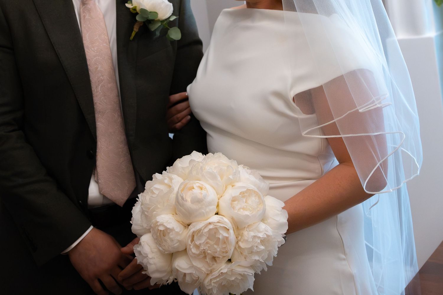Bride in white satin gown holding elegant white peony bouquet stands beside groom in dark suit with pink tie.