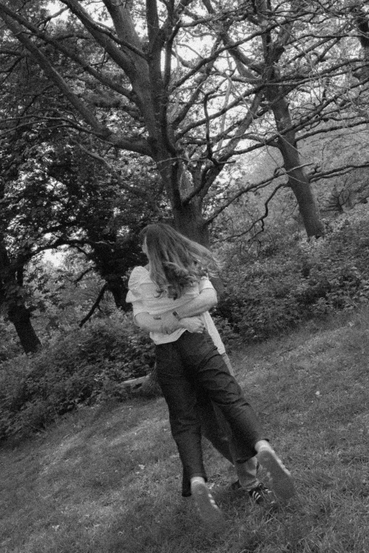 Black and white photo of a woman dancing outdoors in a park surrounded by large trees and lush greenery.