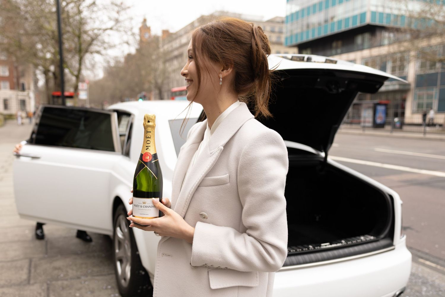 Elegant woman in white blazer holding Moet champagne bottle beside white Rolls-Royce on city street.