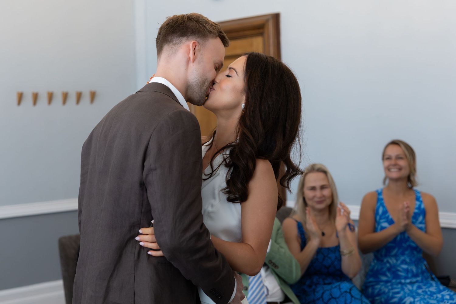 A couple shares their first kiss as newlyweds while wedding guests clap and smile in the background.