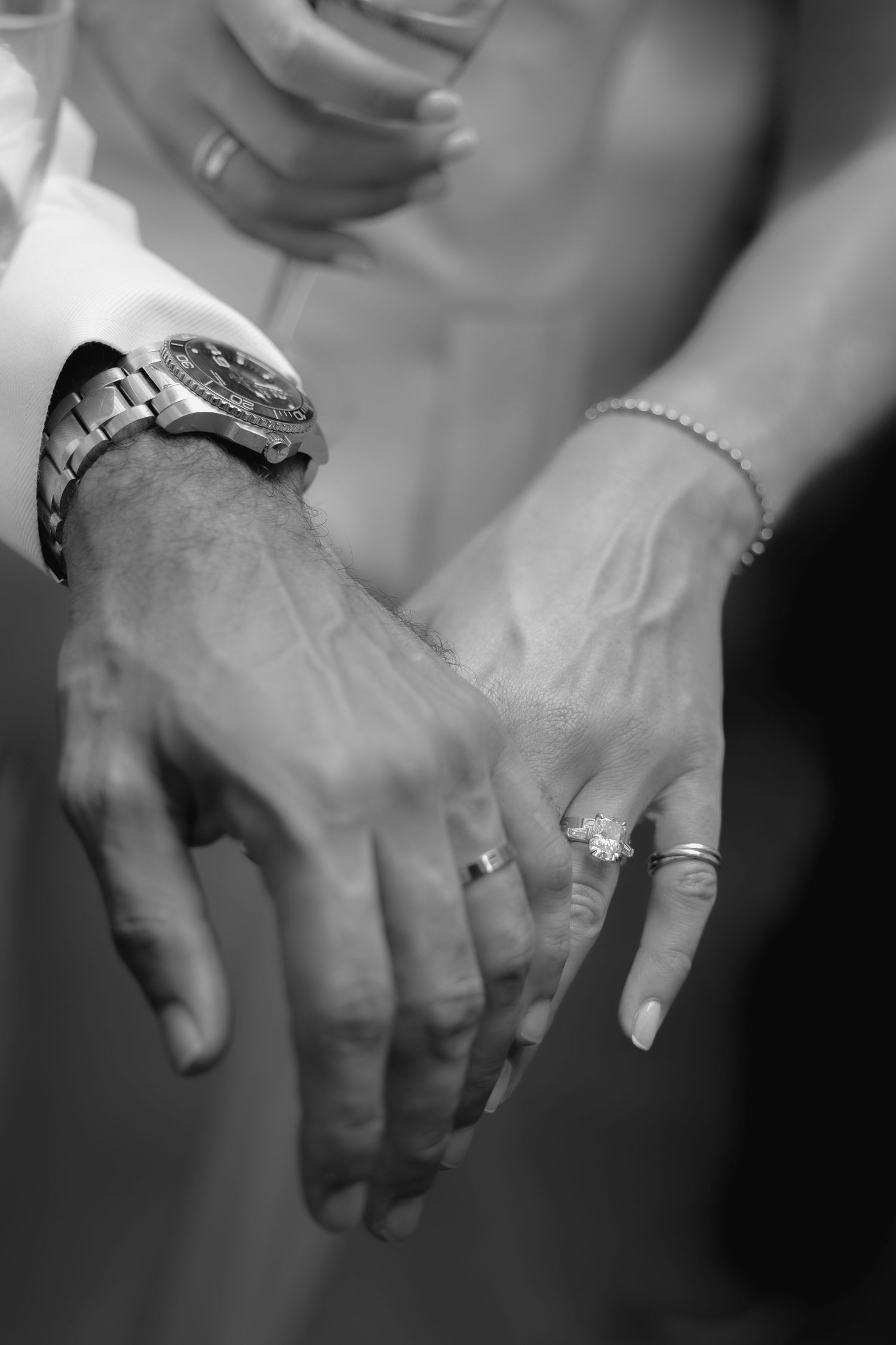 Black and white photo of couple's hands showing wedding rings, groom's watch, and bride's diamond engagement ring.