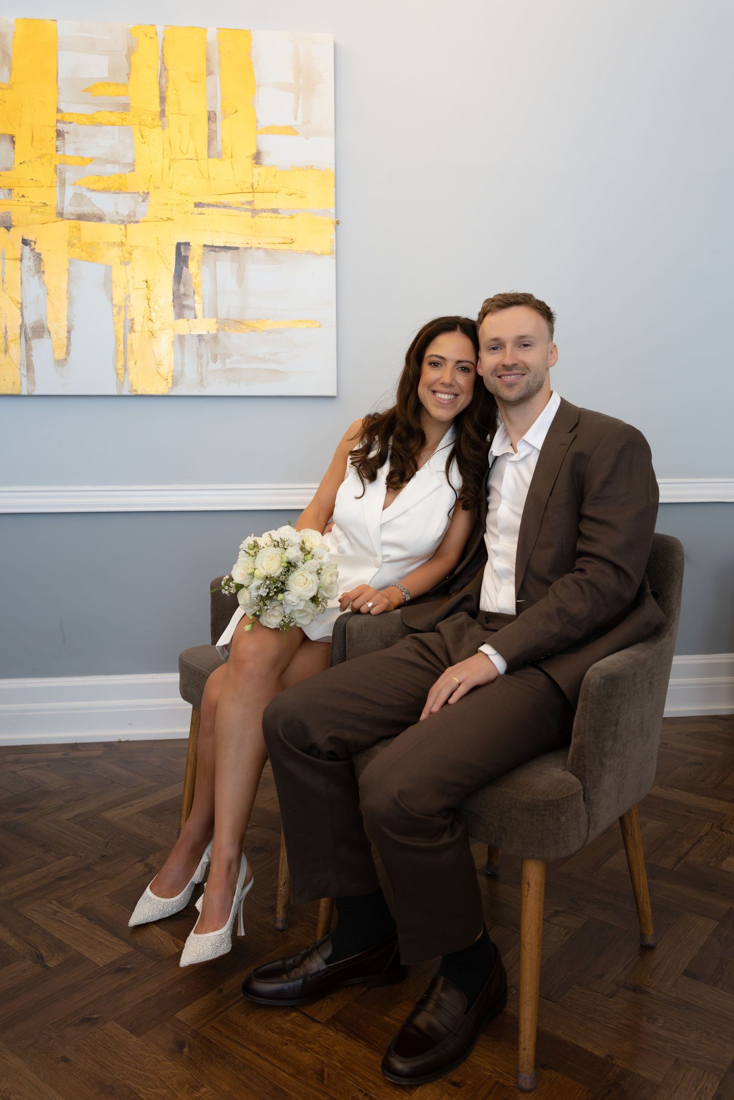 Couple in wedding attire sitting in a gray chair, woman in white dress with bouquet, man in brown suit, yellow abstract art behind.