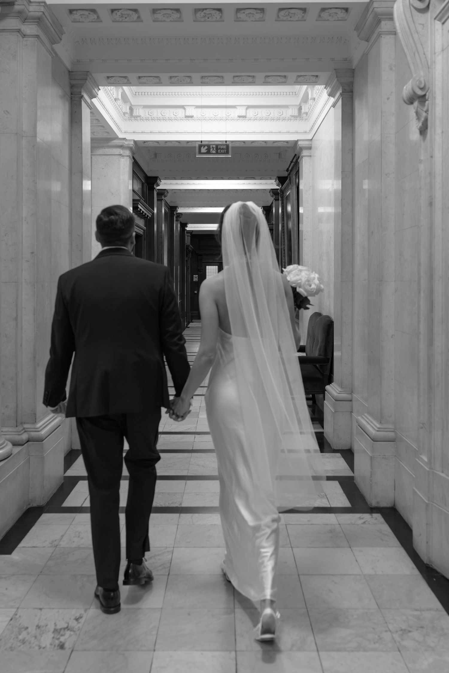 Black and white photo of bride and groom walking hand in hand through marble corridor.