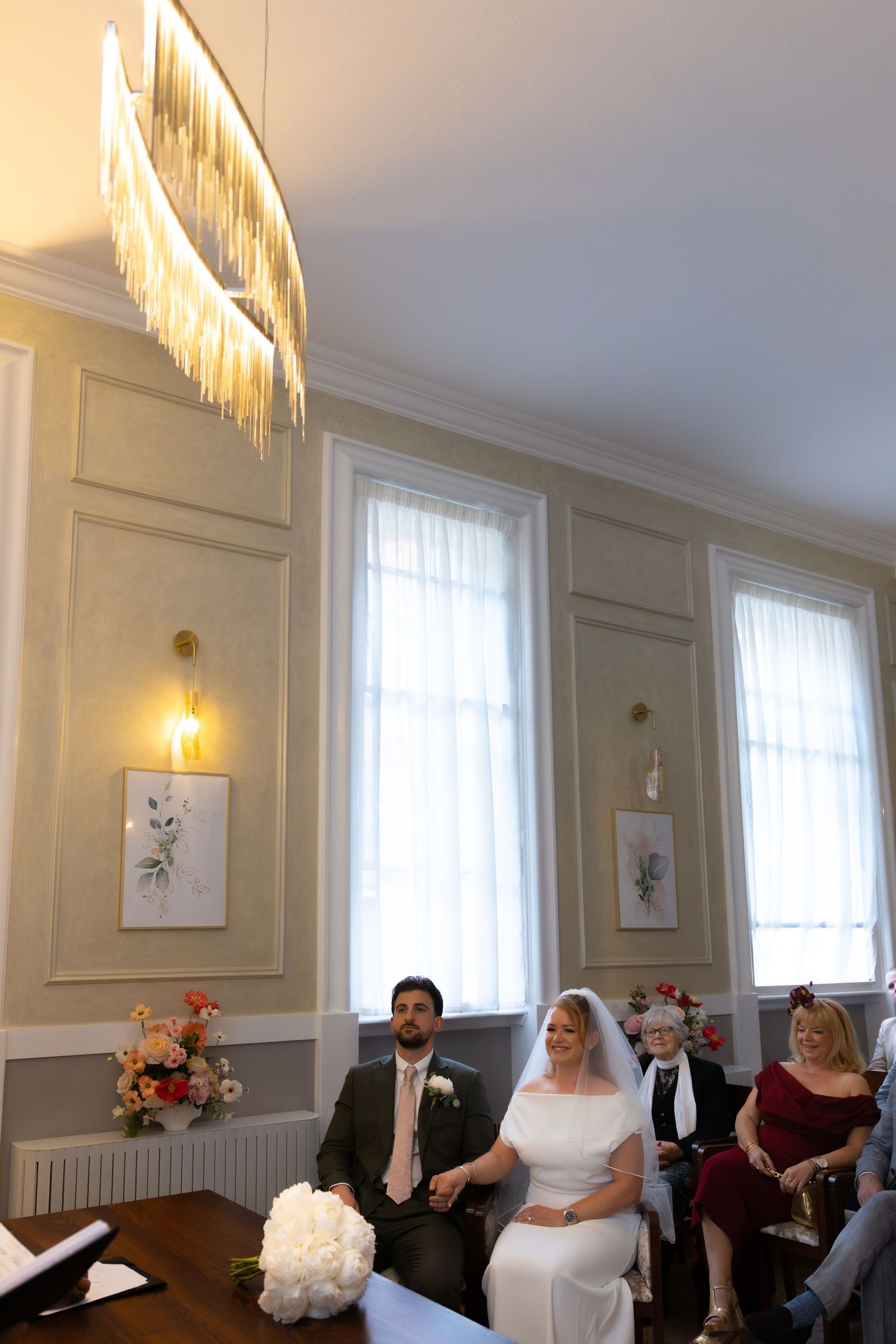Bride and groom seated at civil wedding ceremony in elegant room with chandelier and tall windows.