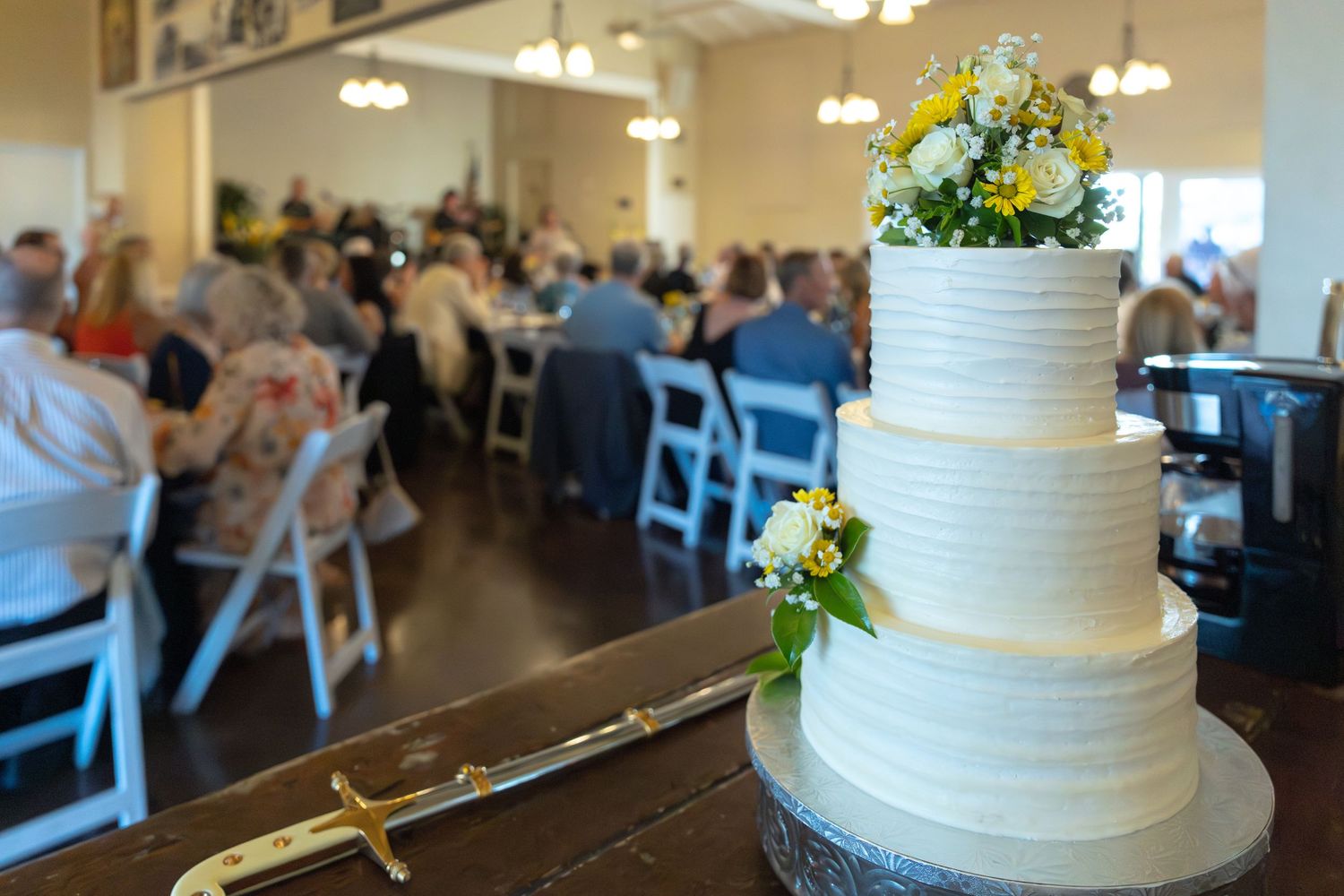 Elegant three-tier white wedding cake with yellow flowers at a reception hall filled with guests.