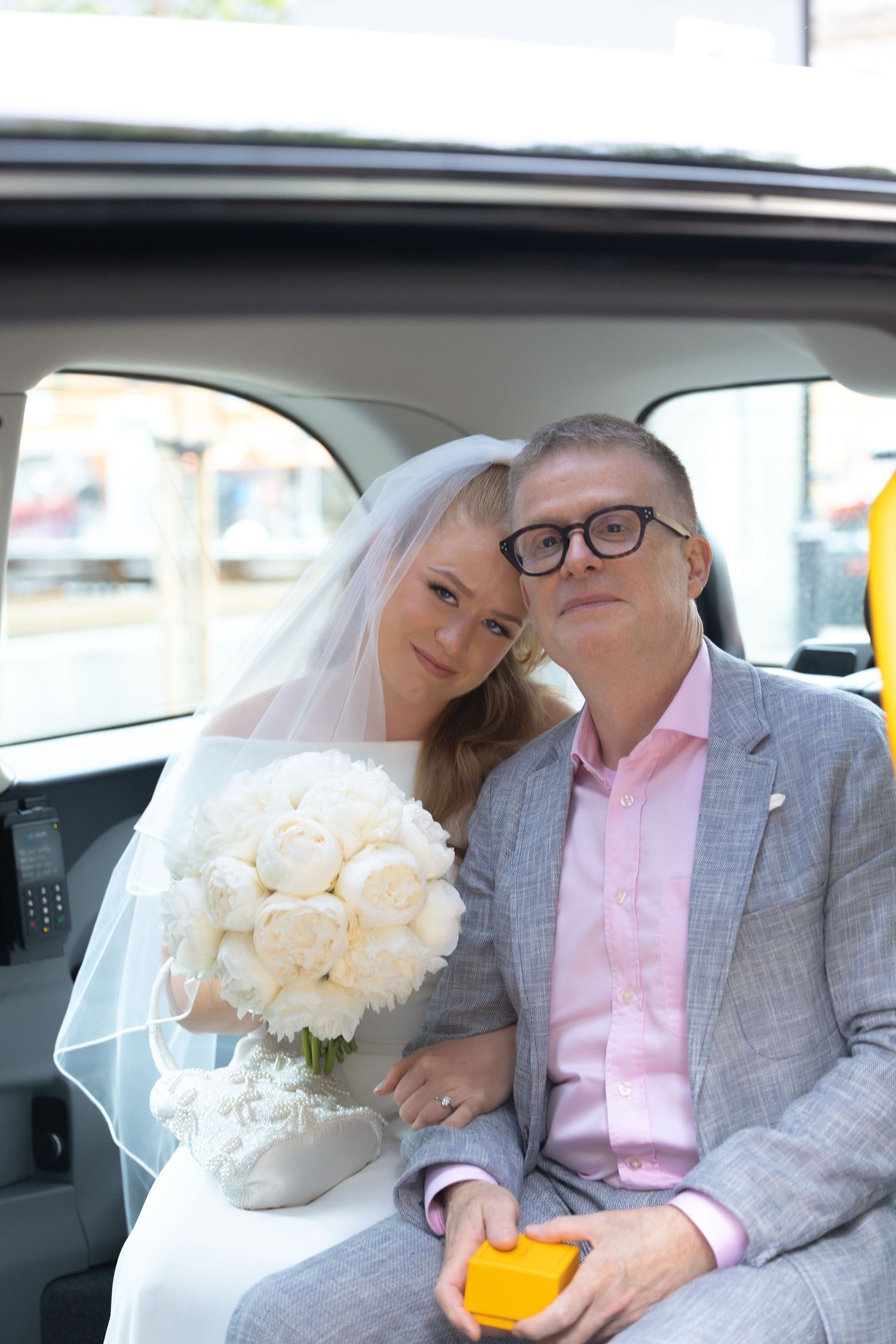 Bride and groom sitting together in a taxi cab, bride holding white peony bouquet wearing veil.