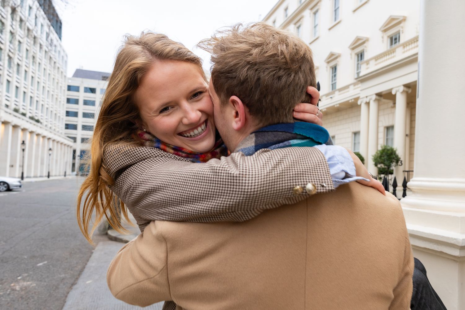 A romantic couple embraces on a city street with classic architecture in the background.