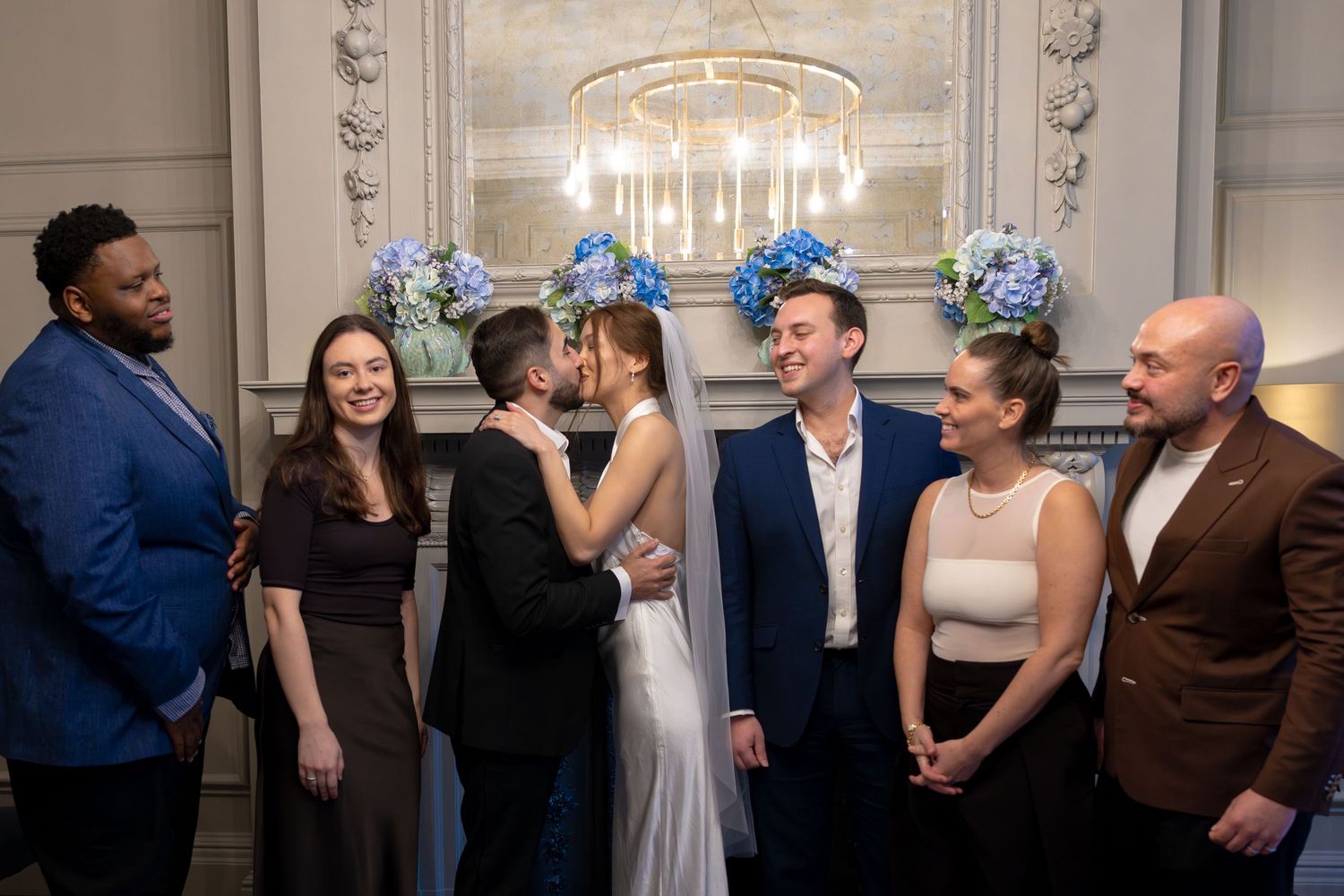 Couple shares first kiss at elegant wedding ceremony surrounded by smiling guests near floral chandelier.