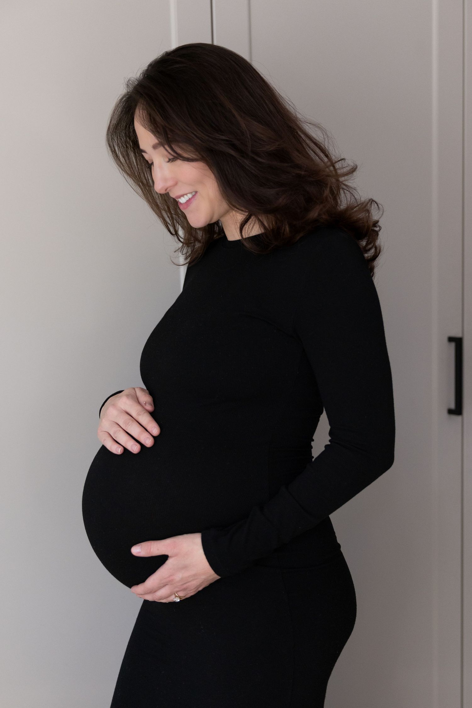 Pregnant woman in black fitted dress cradling baby bump while smiling in bright room.