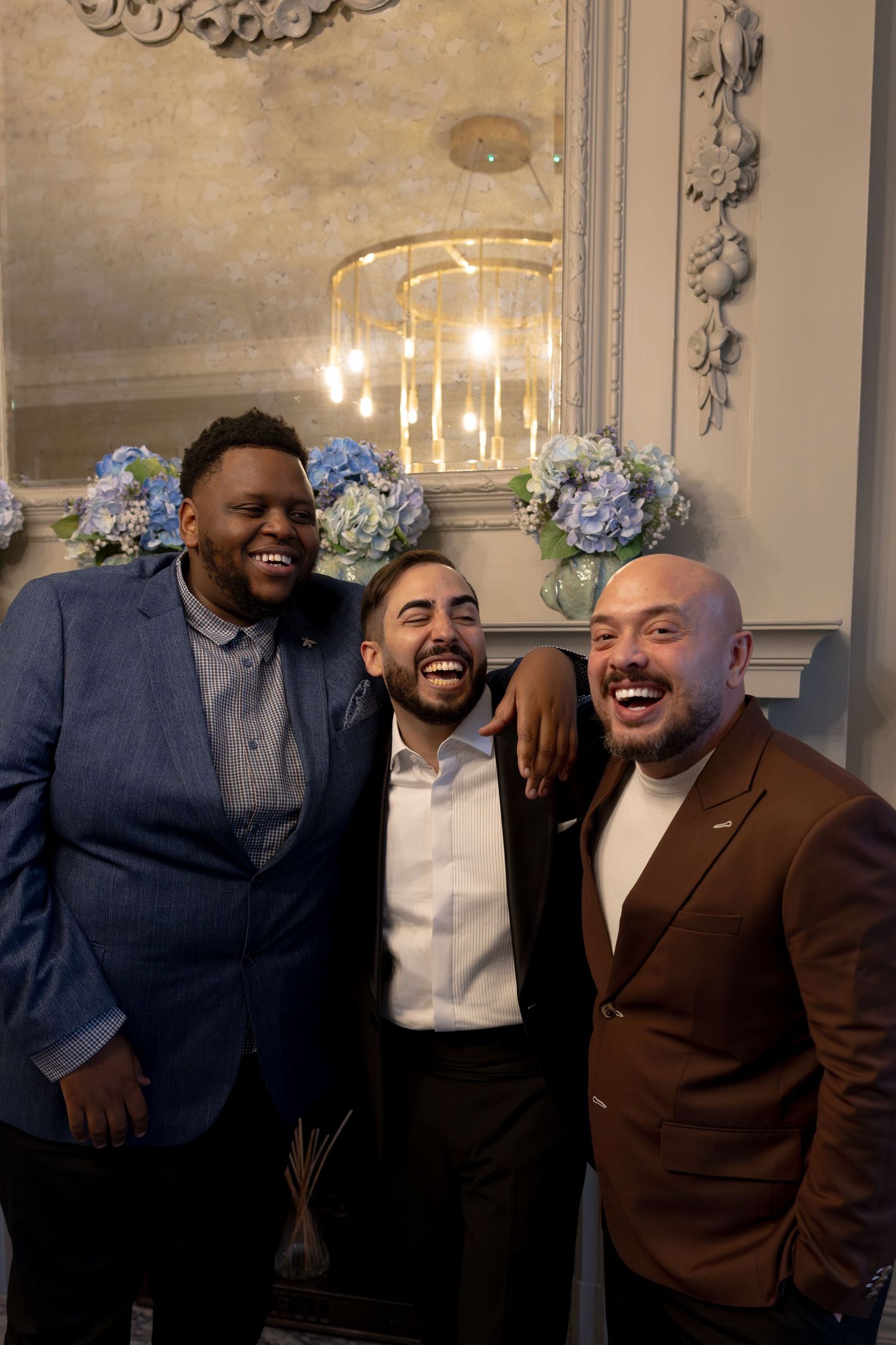 Three men laughing together at wedding reception near decorative fireplace with blue hydrangeas.