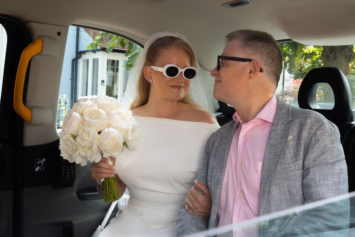 Bride in white dress and sunglasses holds bouquet beside suited man in black taxi cab.