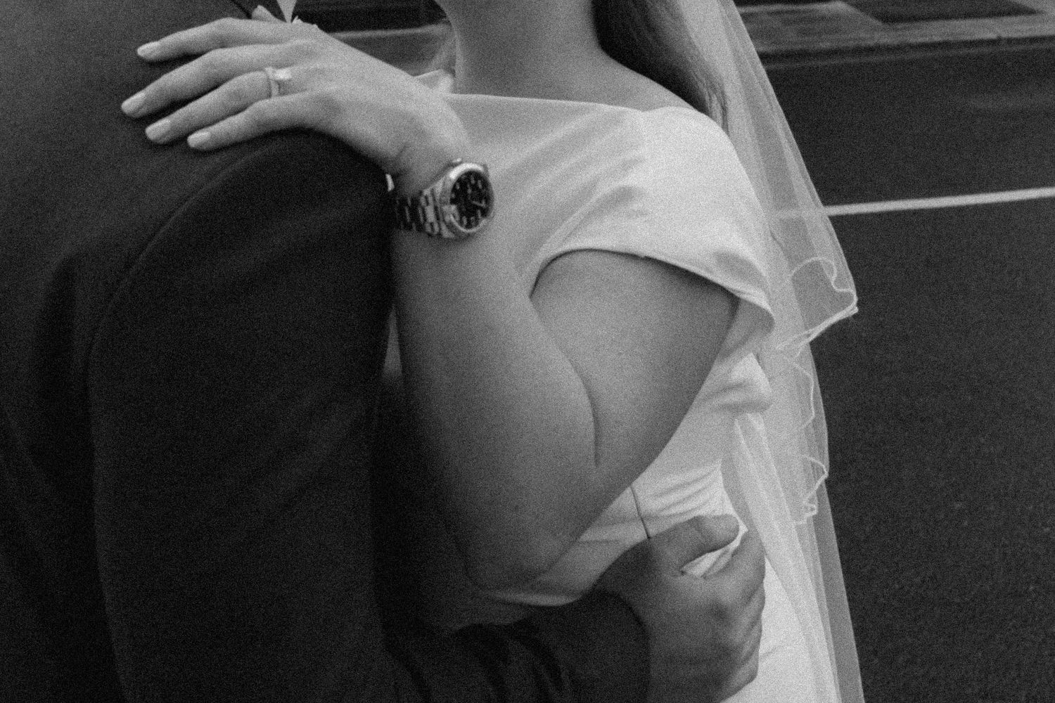 Black and white photo of a bride in white dress and veil embracing a groom, showcasing a silver watch.