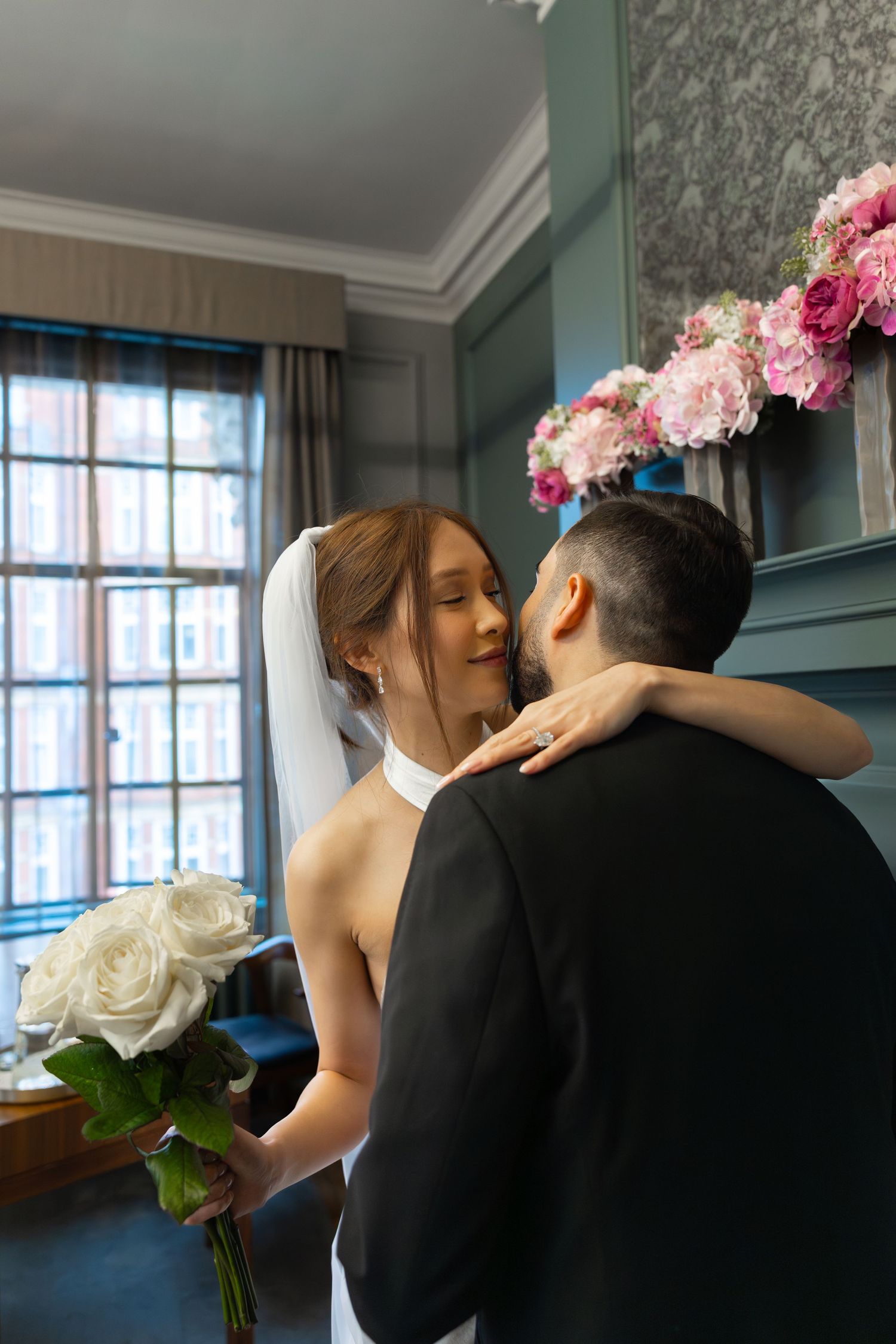 Bride and groom sharing an intimate embrace indoors, bride holding white roses with pink flowers in background.