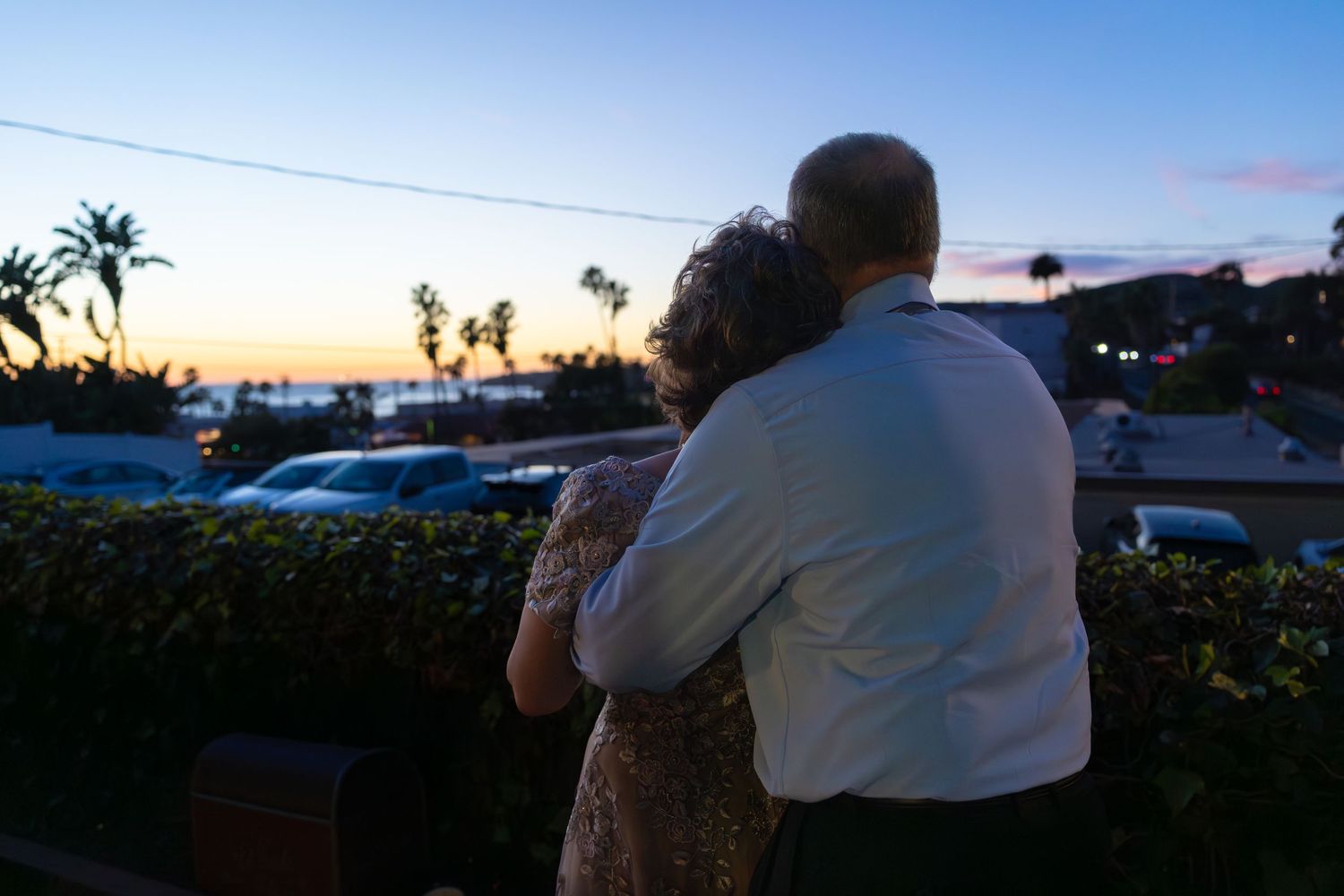 Couple embracing at sunset overlooking a coastal town with palm trees and colorful sky.
