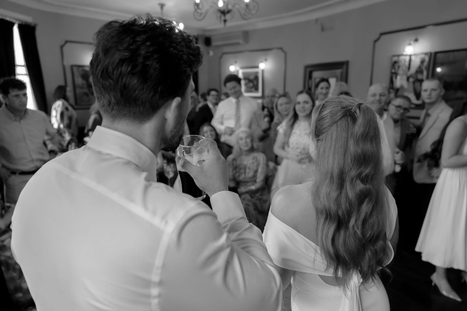 Black and white wedding toast with couple raising glasses to smiling guests in elegant reception hall.