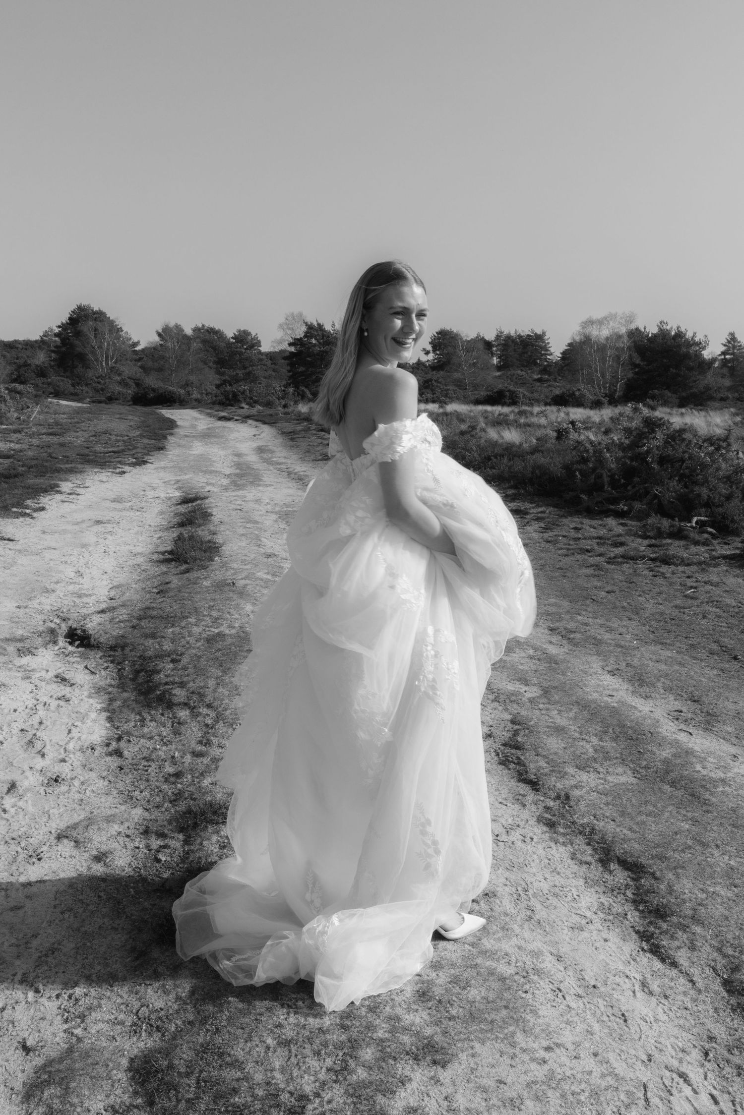Black and white portrait of a smiling bride in an off-shoulder ball gown standing on a rural dirt path outdoors.