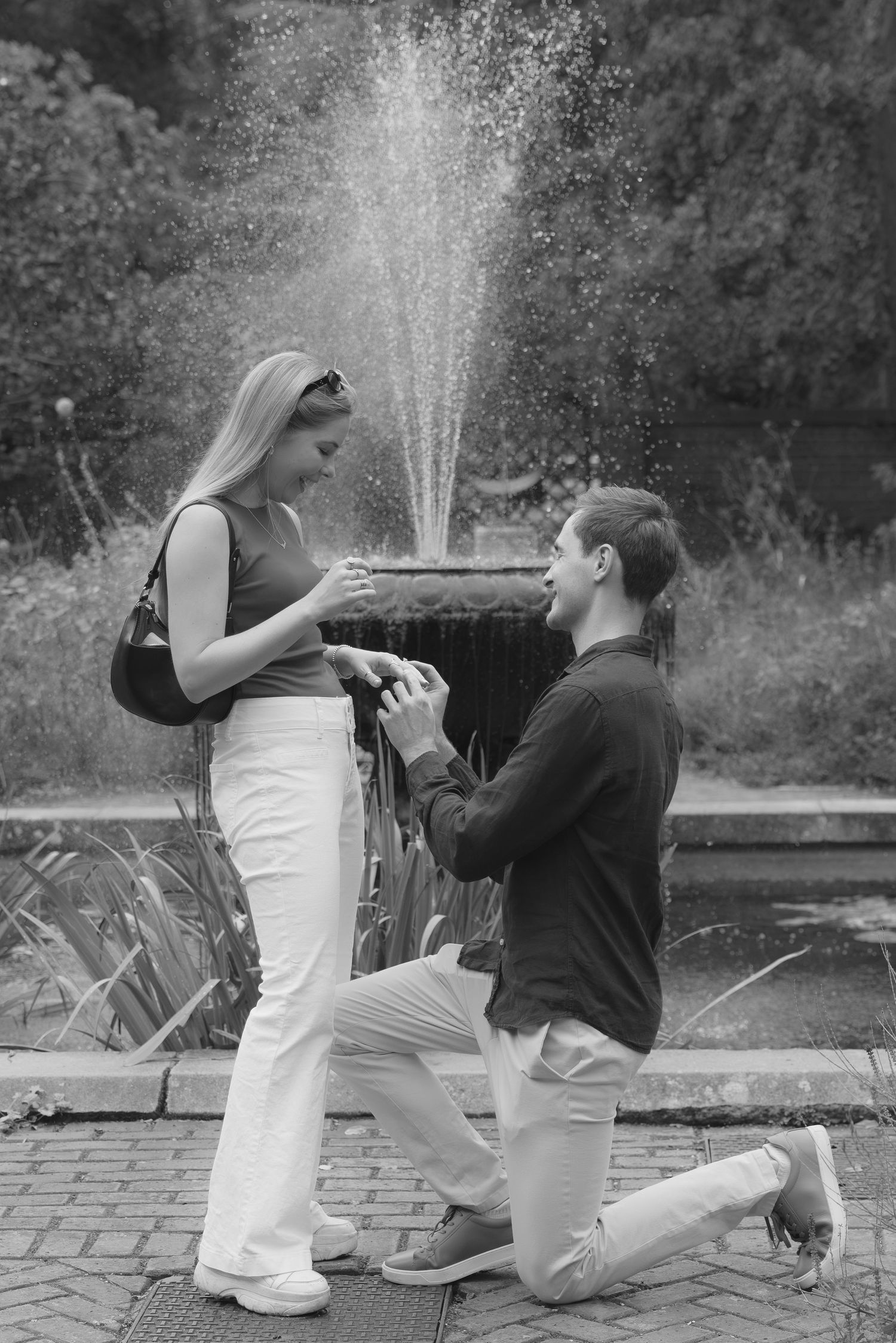 Black and white photo of a man on one knee proposing to a woman in front of a garden fountain.