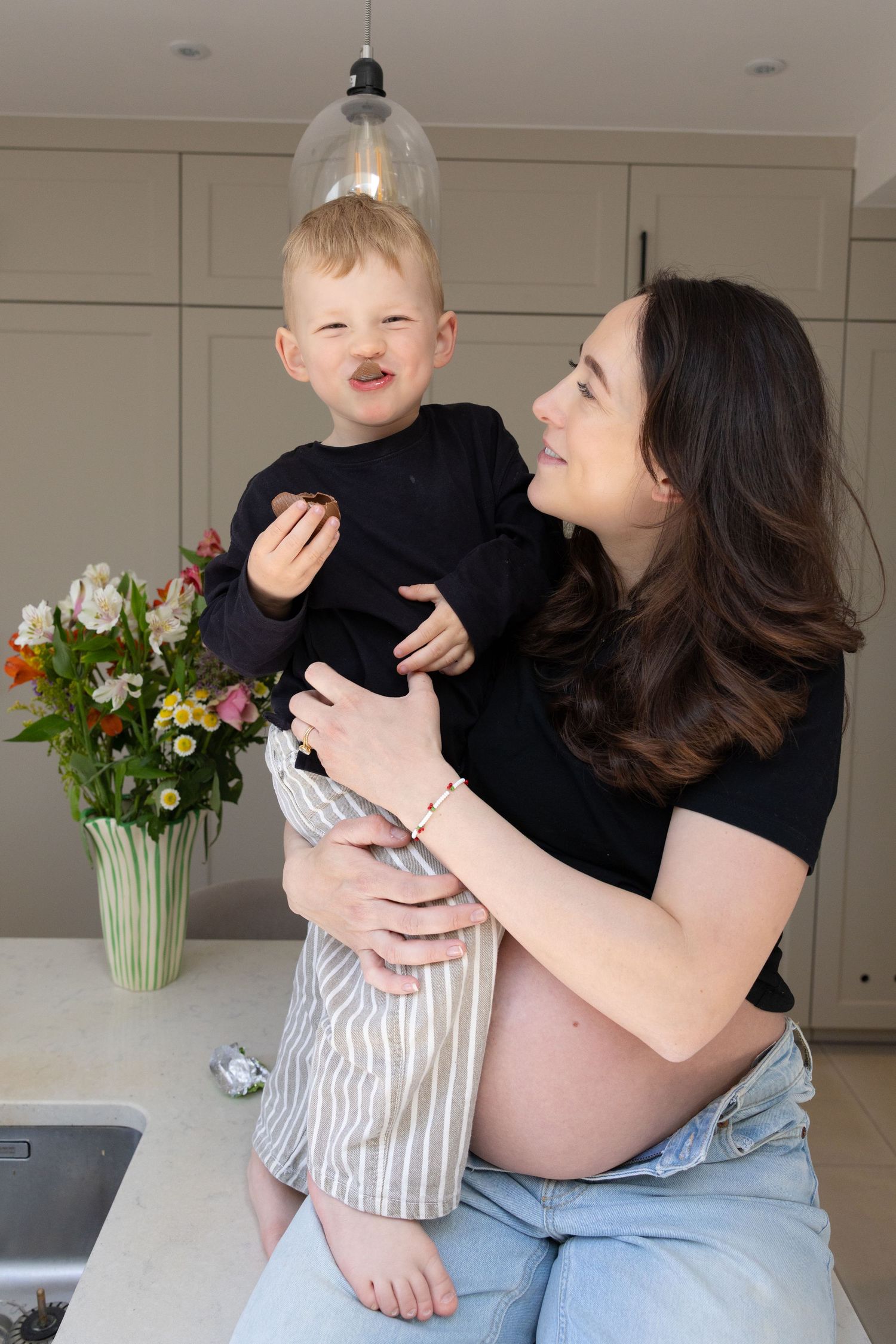 Pregnant mother holding toddler son on kitchen counter, child making funny face.