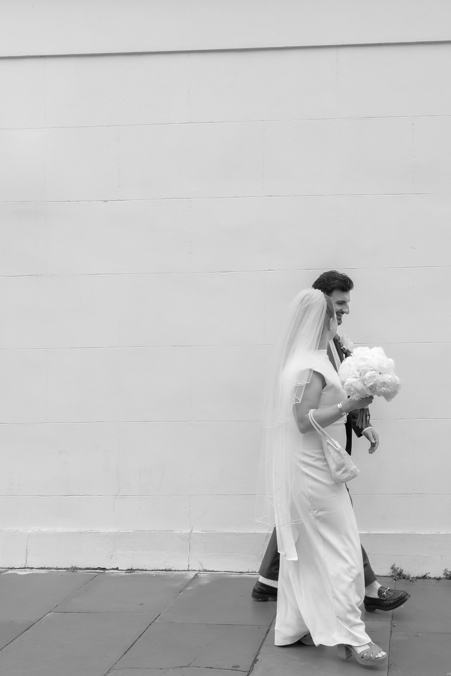 Bride and groom walking past white wall, bride in elegant white dress holding bouquet, black and white.
