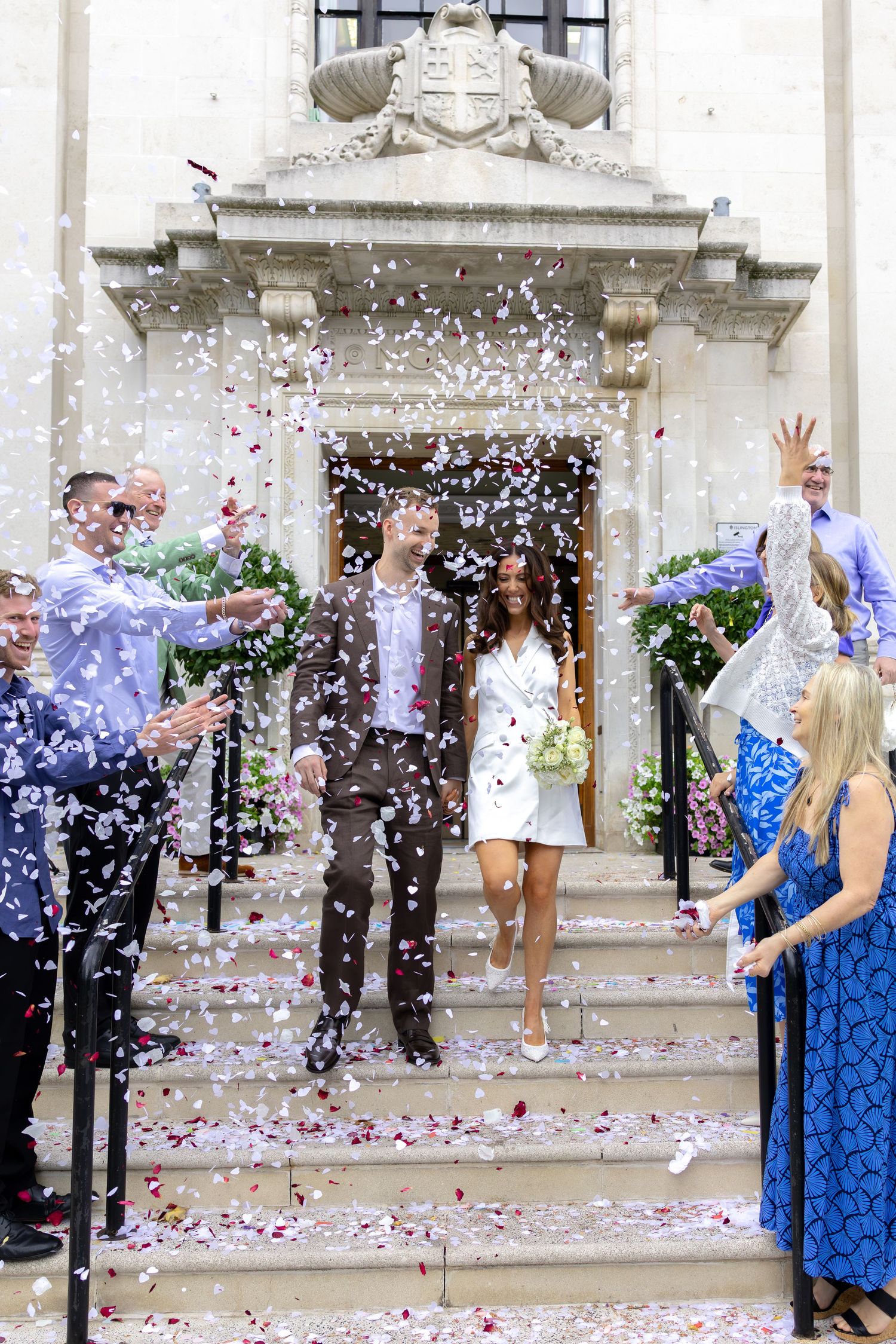 Newlywed couple exits building amid confetti shower, surrounded by cheering guests on steps.
