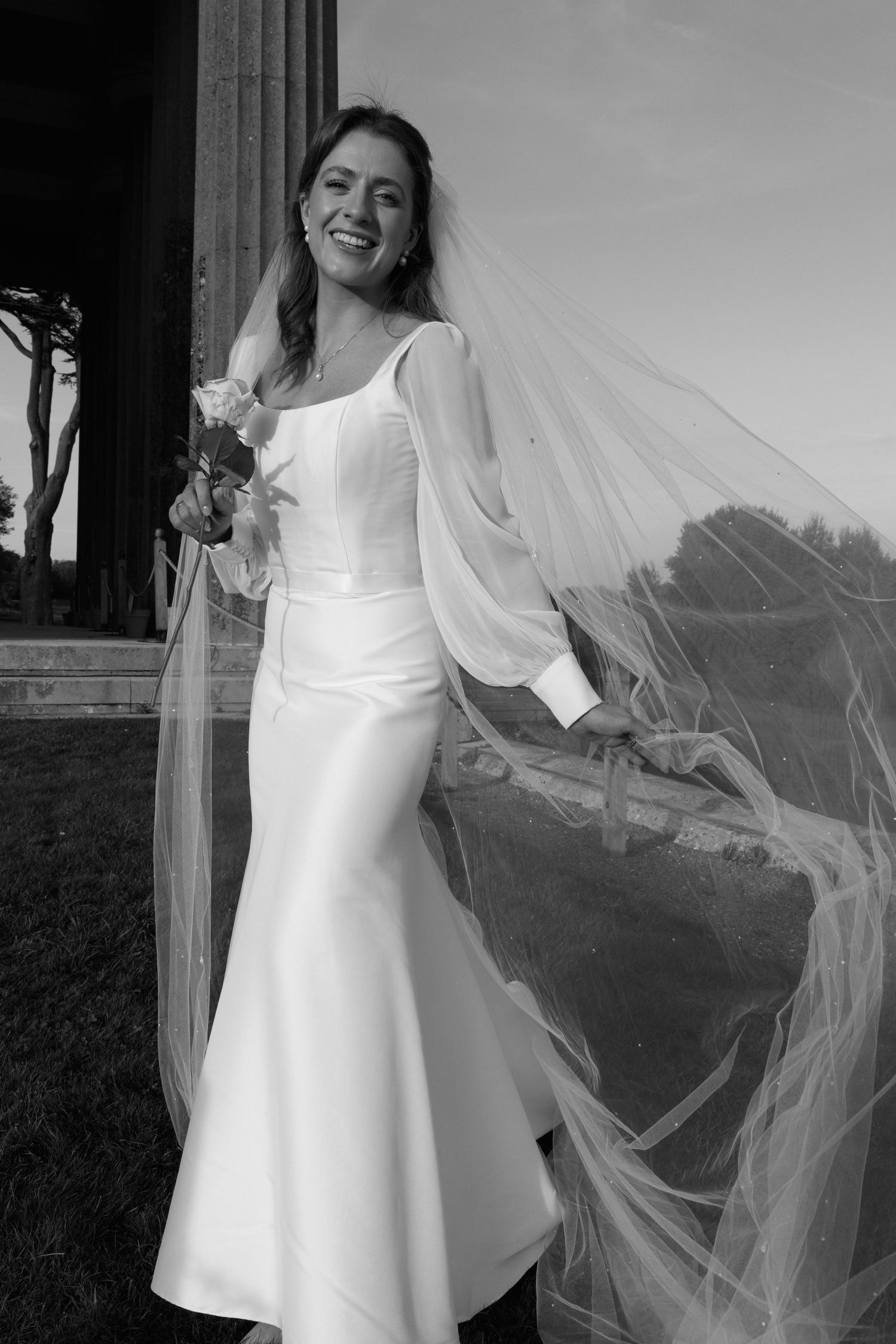 A black and white portrait of a bride in a flowing dress with sheer veil holding flowers outdoors.