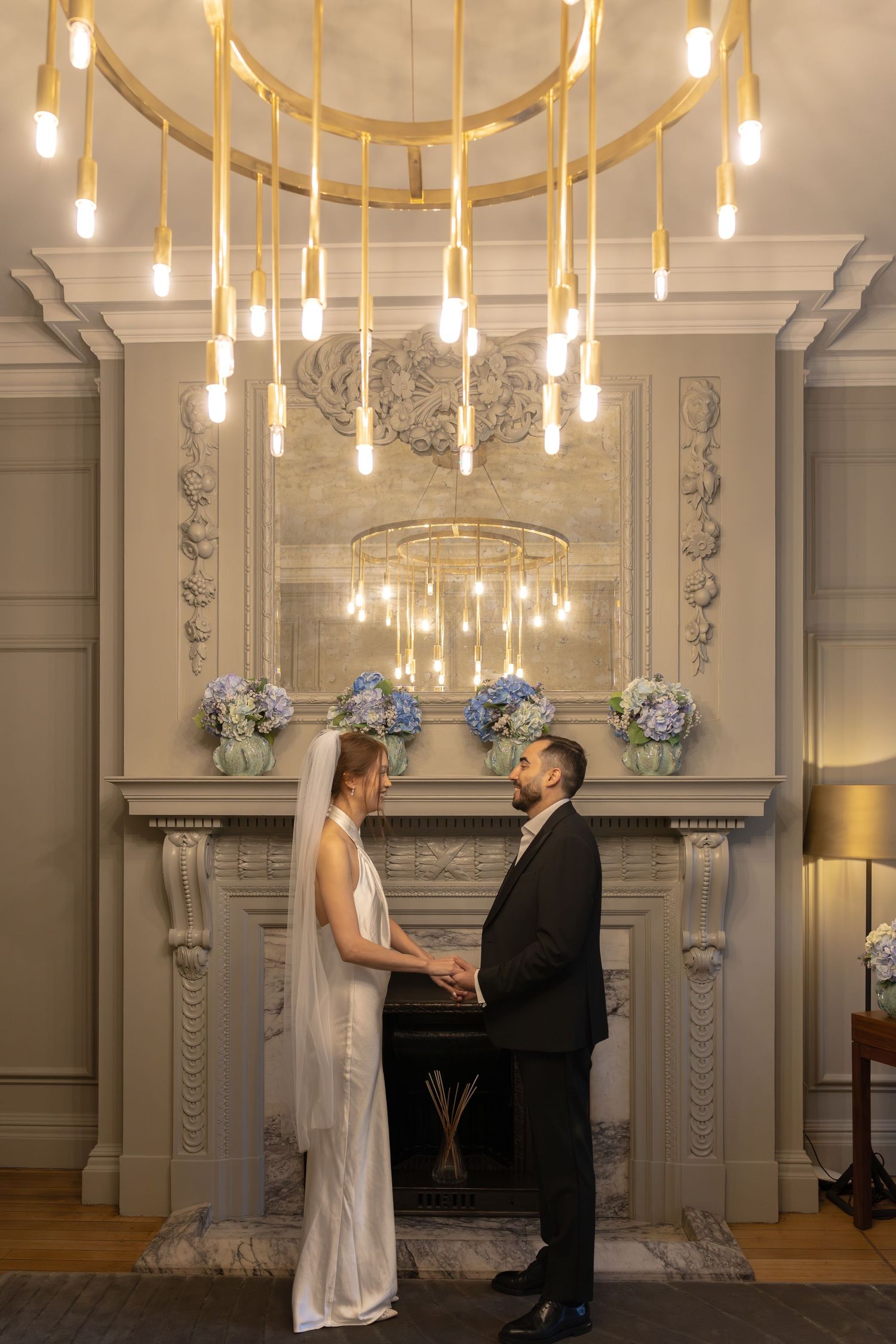 Bride and groom hold hands before ornate fireplace under elegant gold chandelier with Edison bulbs.