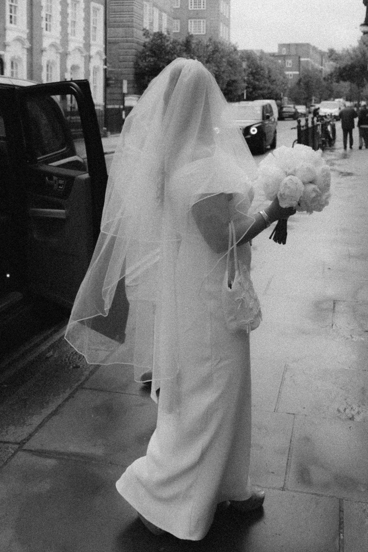 Bride in white gown and veil exits a black car on a rainy wedding day in London.
