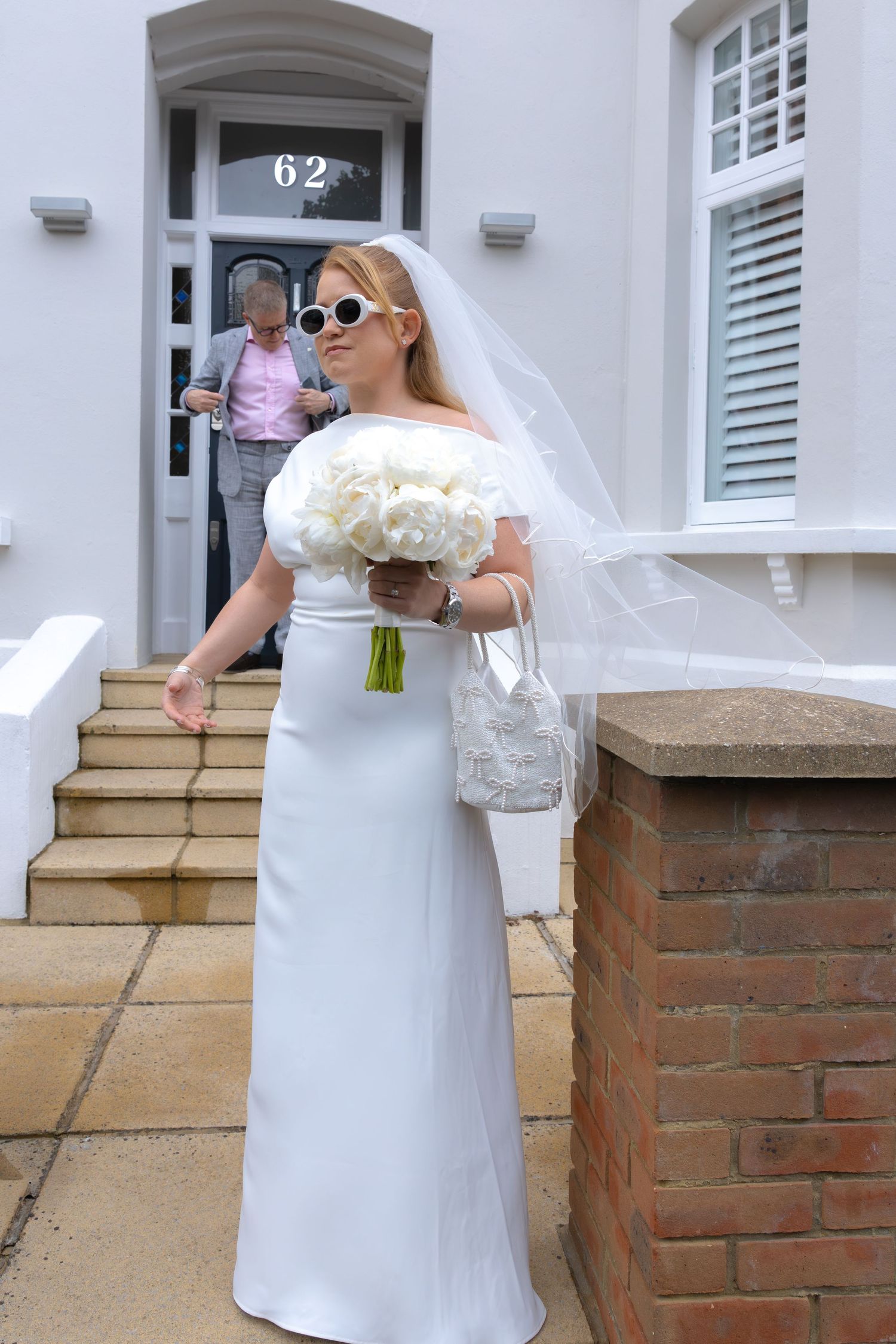 Bride in white off-shoulder gown, veil, sunglasses, holding white peony bouquet outside house number 62.
