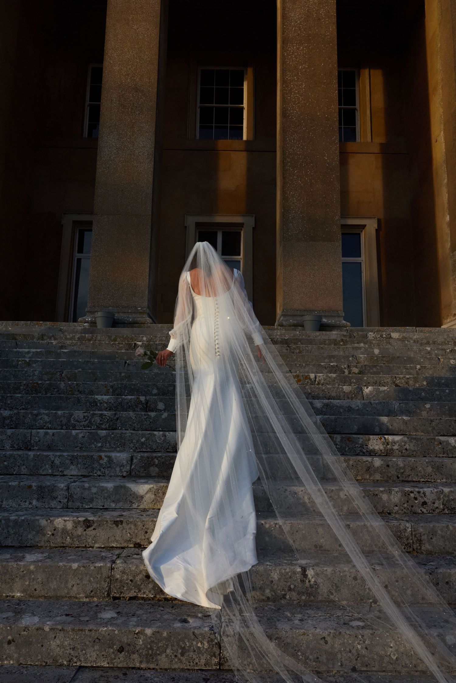 A bride in a flowing dress and veil ascends stone steps at dusk, captured from behind.