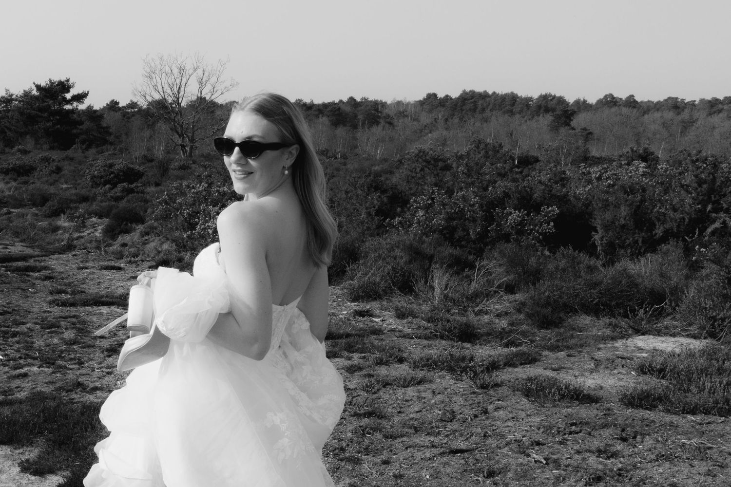 Woman in white gown and sunglasses poses in black and white outdoor landscape with shrubs.