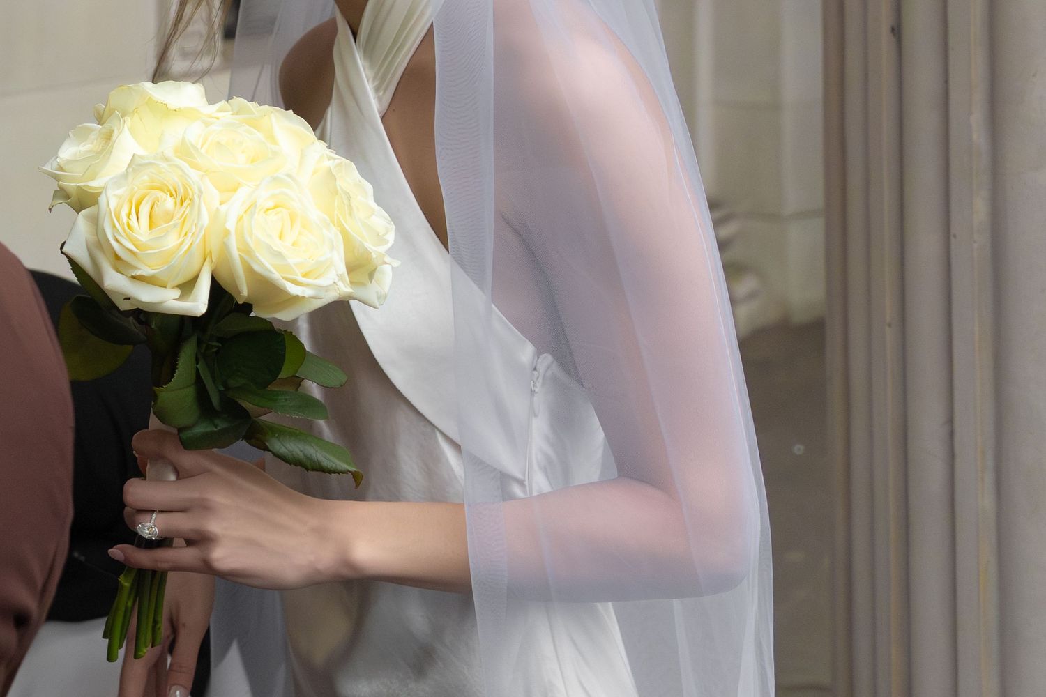 Bride in white wedding dress holding bouquet of cream roses with veil, elegant bridal detail shot.