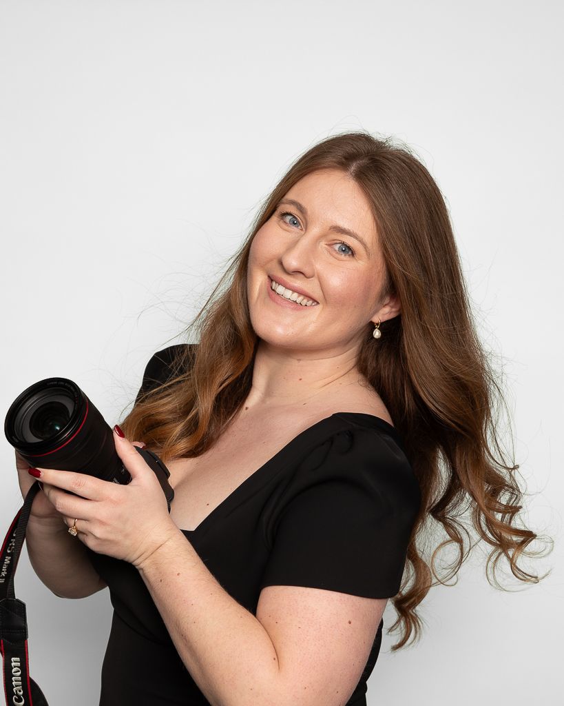 Photographer in black holding a camera and posing against white background.