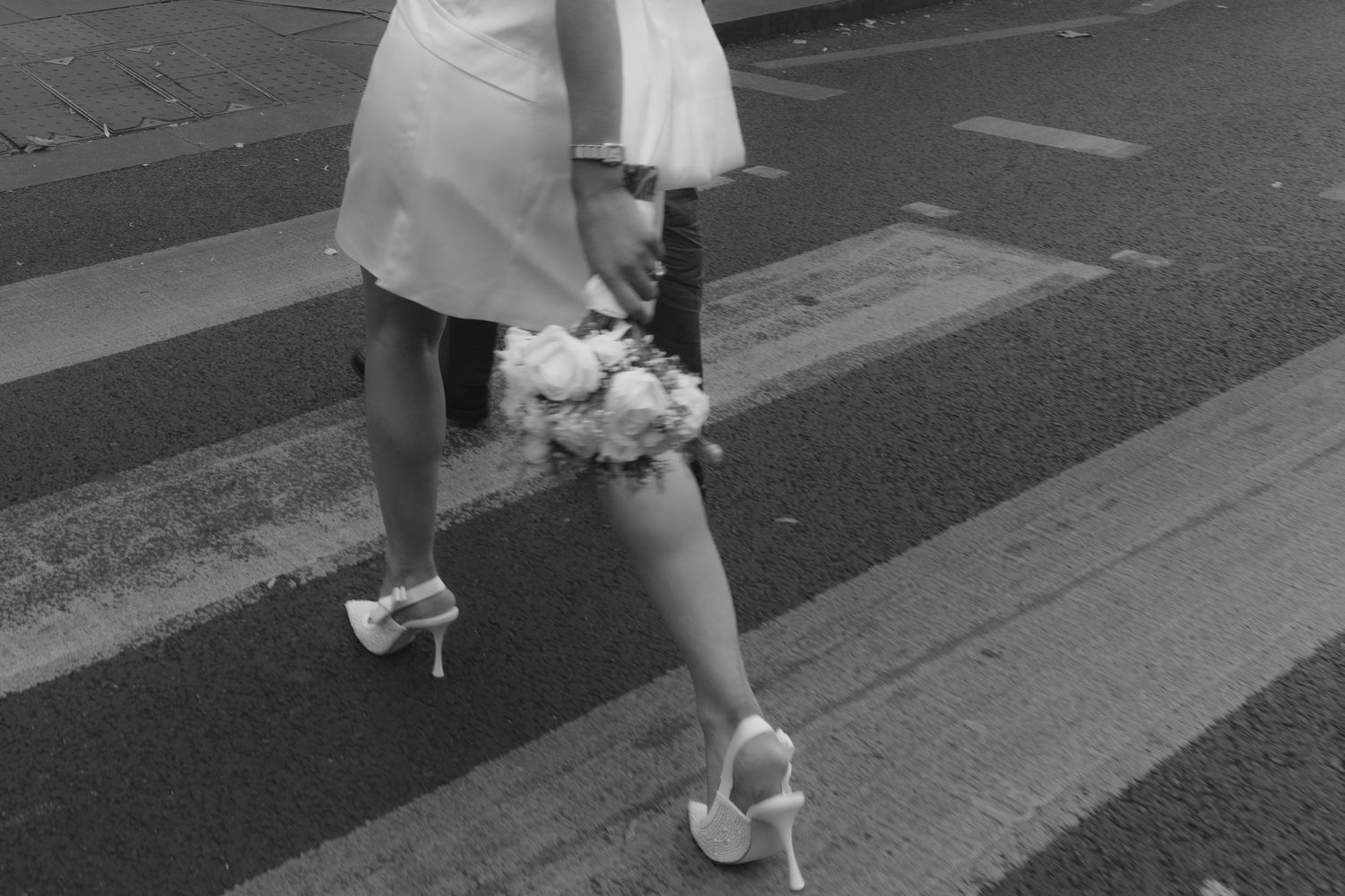 Black and white photo of a bride in white dress and heels crossing a zebra crossing while holding a bouquet.