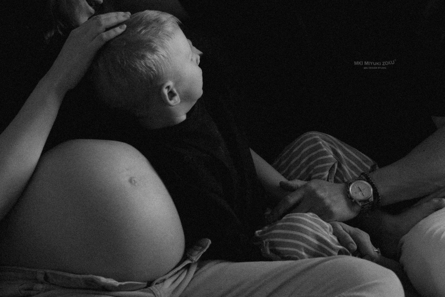 Black and white photo of a child resting against a pregnant womans belly indoors.