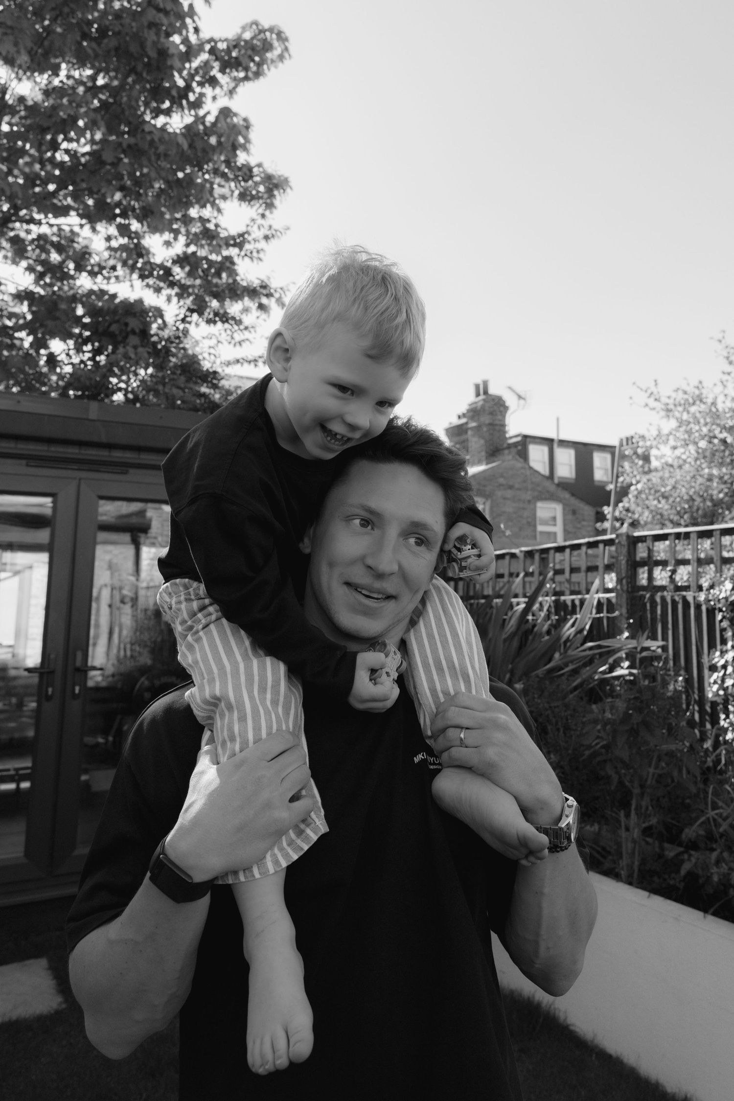 Man smiling with young child on his shoulders in a backyard, black and white photo.